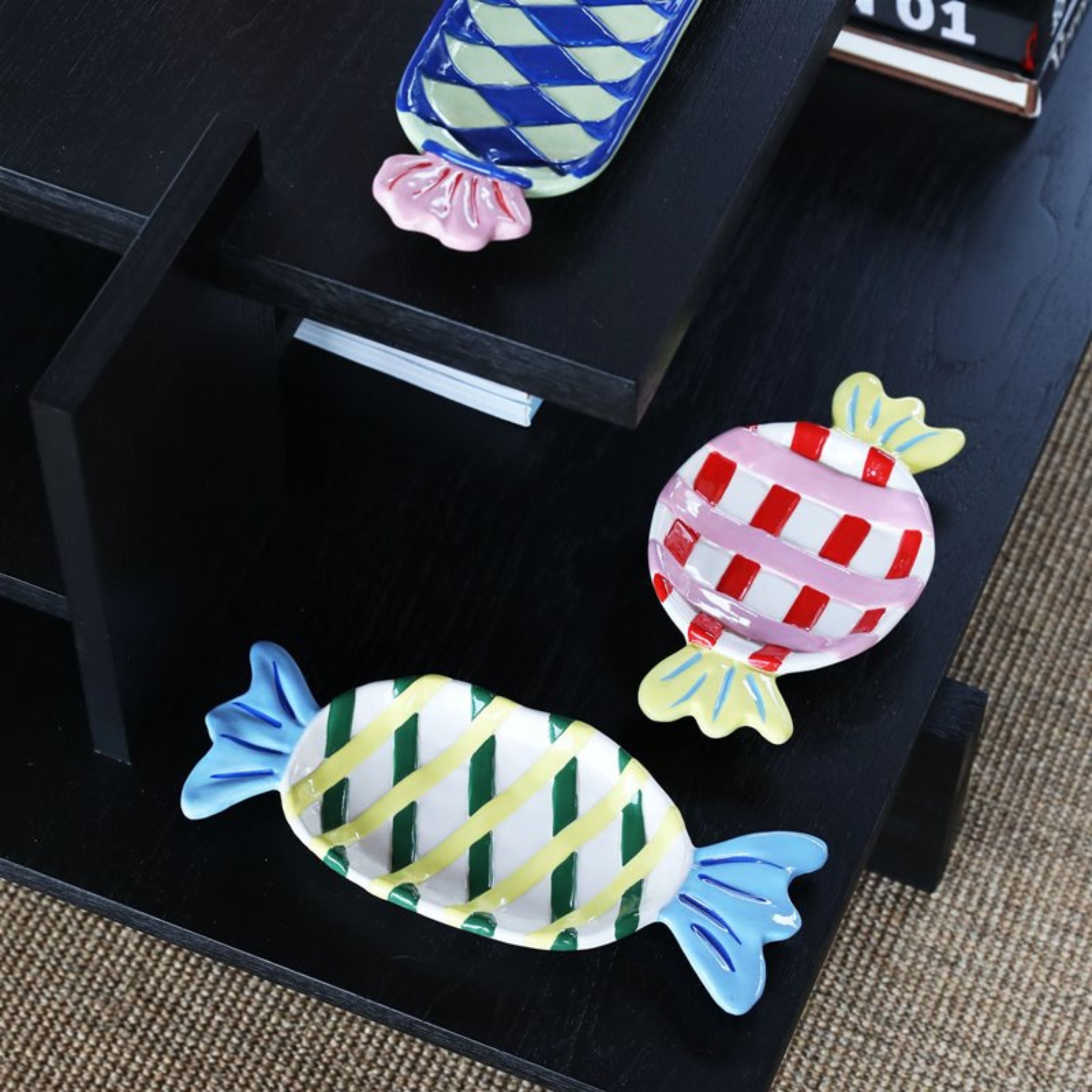 Drie &Klevering Goody Bord 27/11 schaaltjes van keramiek, in de vorm van ingepakte snoepjes met blauwe, roze en gele ruitjespatronen, staan op een zwarte plank boven een geweven vloerkleed.