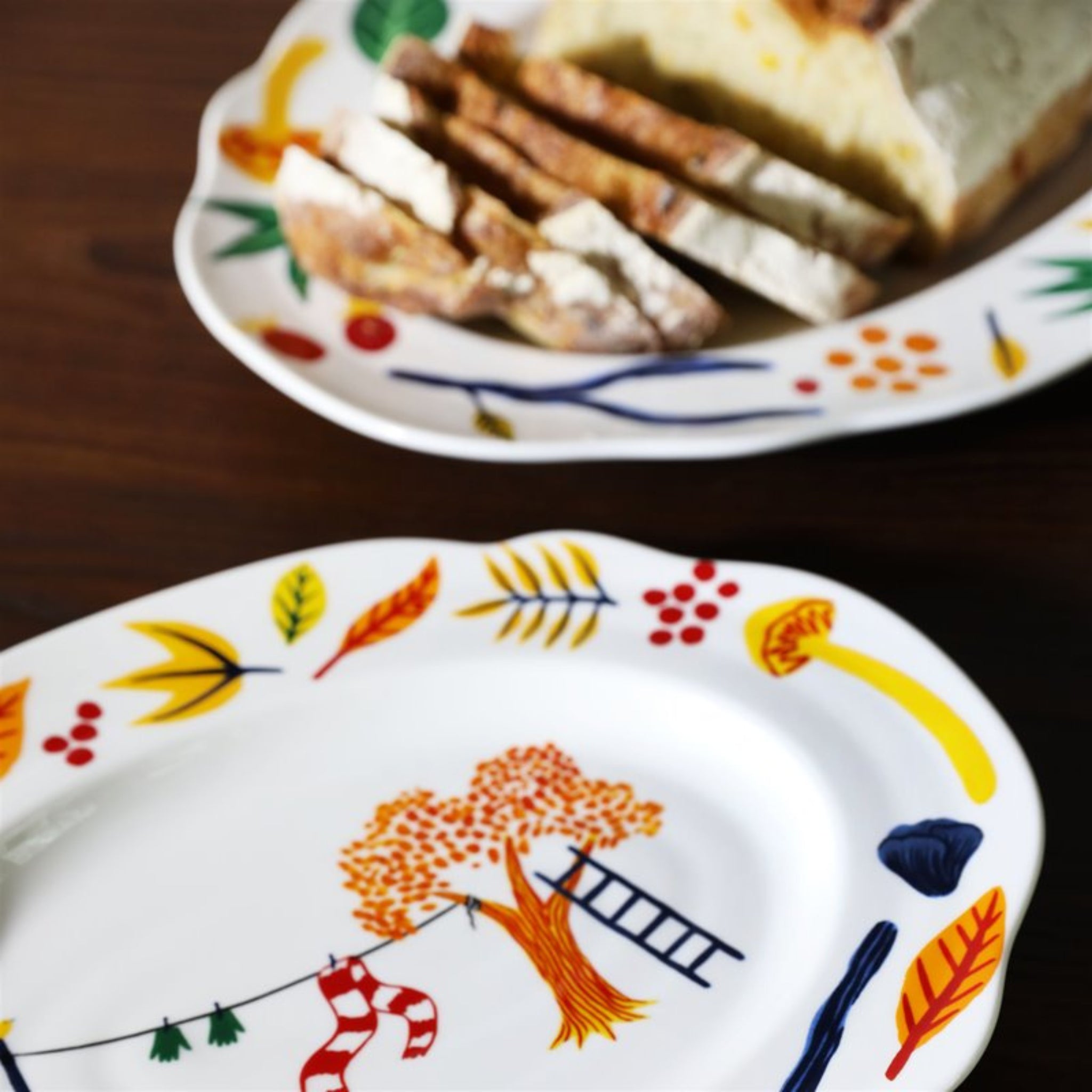 Een close-up van twee &Klevering Haines Schotel 28/22 schotels van &Klevering, een met gesneden brood en een leeg, beide versierd met kleurrijke, grillige illustraties - een ideaal uniek cadeau op een donkere houten tafel.