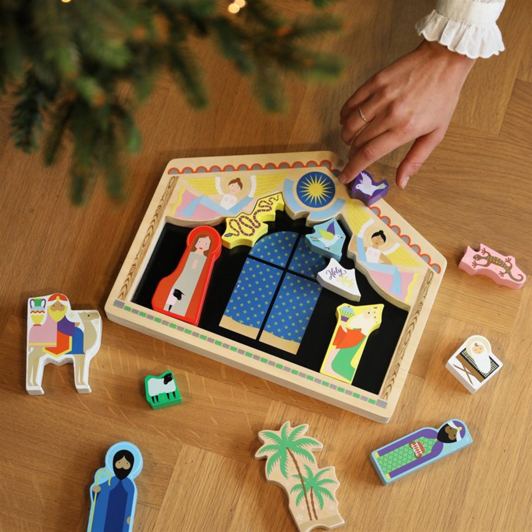 Een hand schikt houten kerststalfiguren uit de &Klevering Kerststal Puzzel van &Klevering op een houten vloer, met Maria, Jozef, kindje Jezus, dieren, een versierde stal en een kerstboom ook zichtbaar.