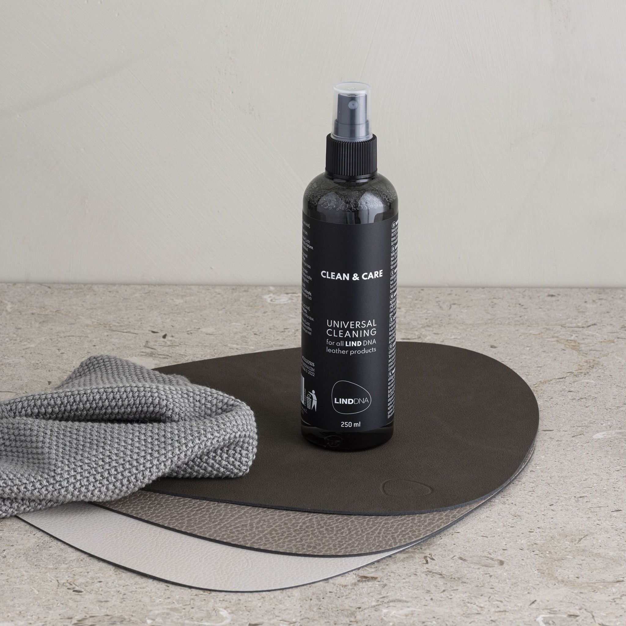 Een zwarte sprayflacon met het label Lind DNA Reinigen & Onderhouden 250 ml van Lind DNA staat op twee gerecyclede leren placemats, met een gevouwen grijs schoonmaakdoekje ernaast, alles op een licht oppervlak.