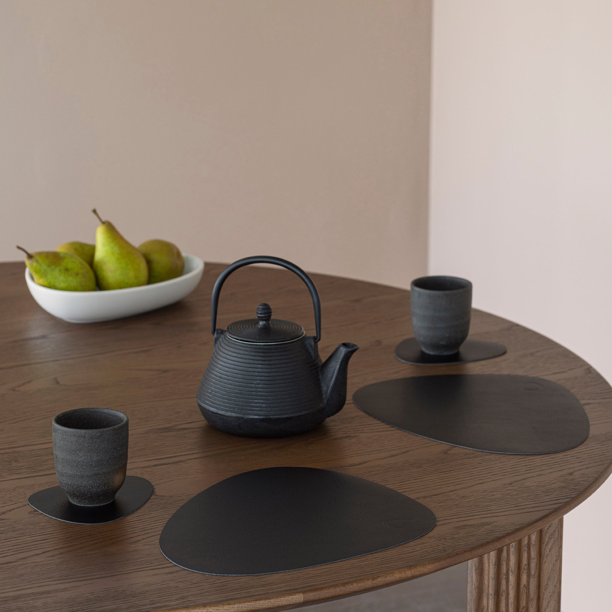 Een ronde houten tafel is gedekt met een zwarte theepot, twee kopjes op schoteltjes en Lind DNA Serene Onderzetter Curve Ash 11/13 onderzetters. Een witte schaal met groene peren staat op de achtergrond en creëert een minimale en moderne look.