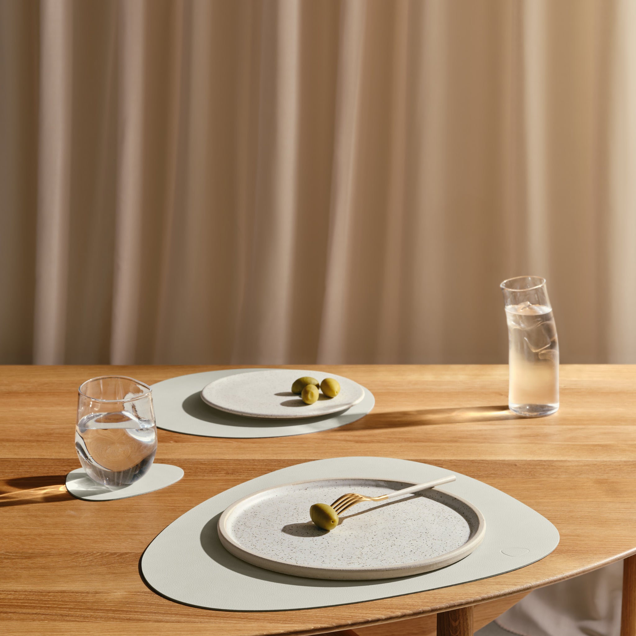 Op een minimalistische tafel staan twee borden met groene olijven en vorken, een glas water en een karaf op onderzetters van Lind DNA Serene Onderzetter Curve Ash 11/13, allemaal op een houten tafel tegen een achtergrond van zacht verlichte gordijnen.