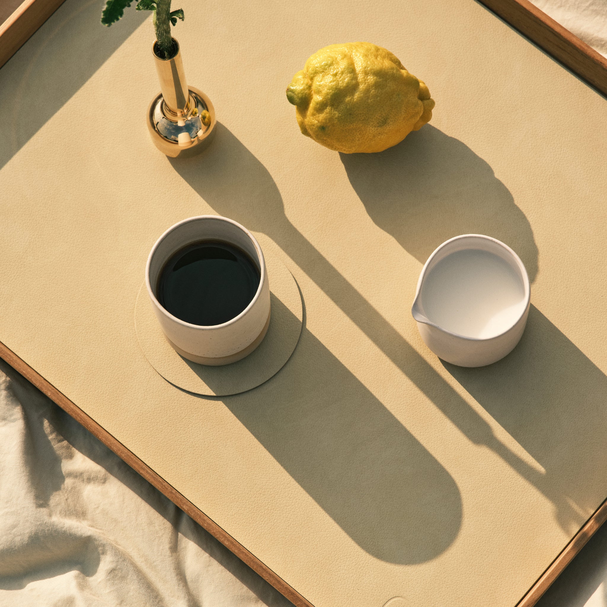 Een kop zwarte koffie, melk, een citroen en een enkele bloem in een vaas liggen op een beige dienblad bovenop Lind DNA Serene Onderzetter Rond Hazel D10 placemats, die allemaal lange schaduwen werpen in het warme zonlicht.