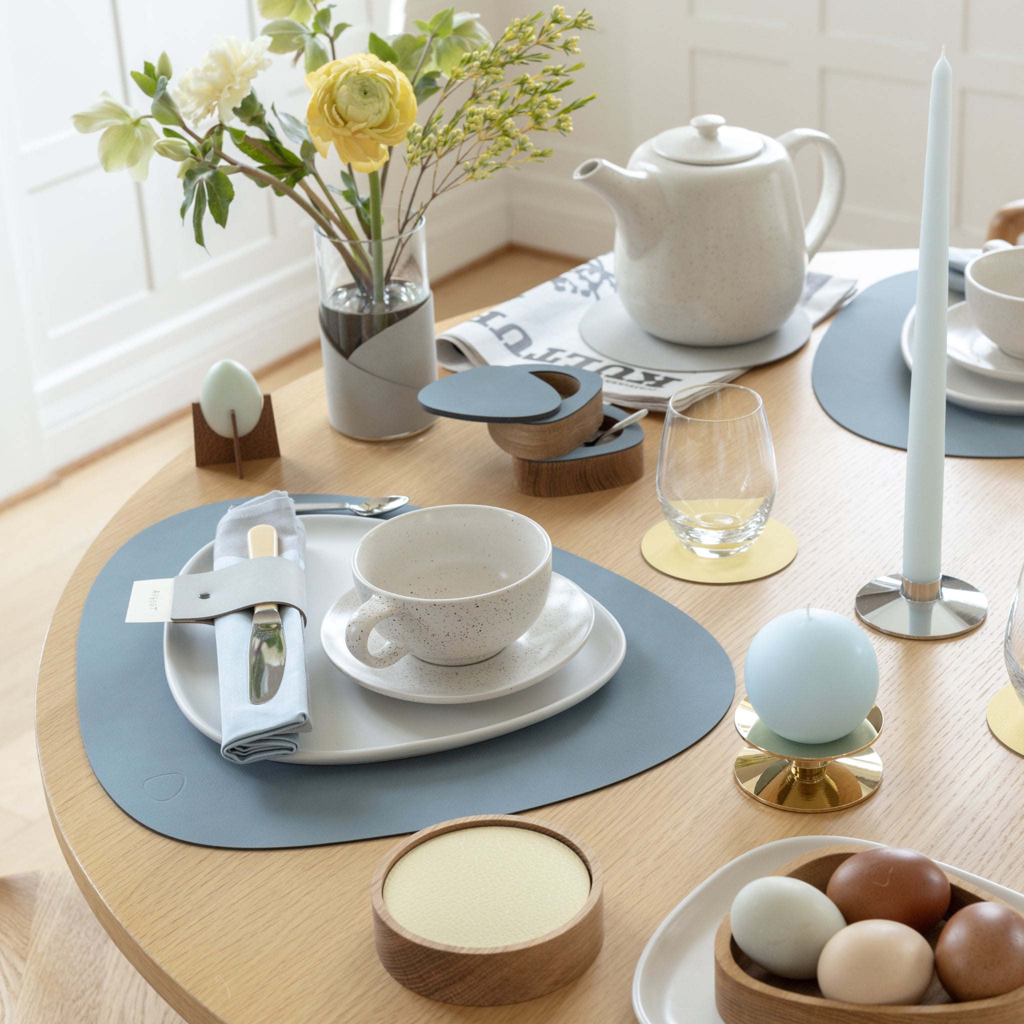 Een moderne houten theetafel is gedekt met witte borden, een theepot, eieren, een pastelblauwe kaars, bloemen in een vaas en onderzetters Serene Onderzetter Rond Hazel D10 van Lind DNA in zachte kleuren. Natuurlijk daglicht verlicht het tafereel.