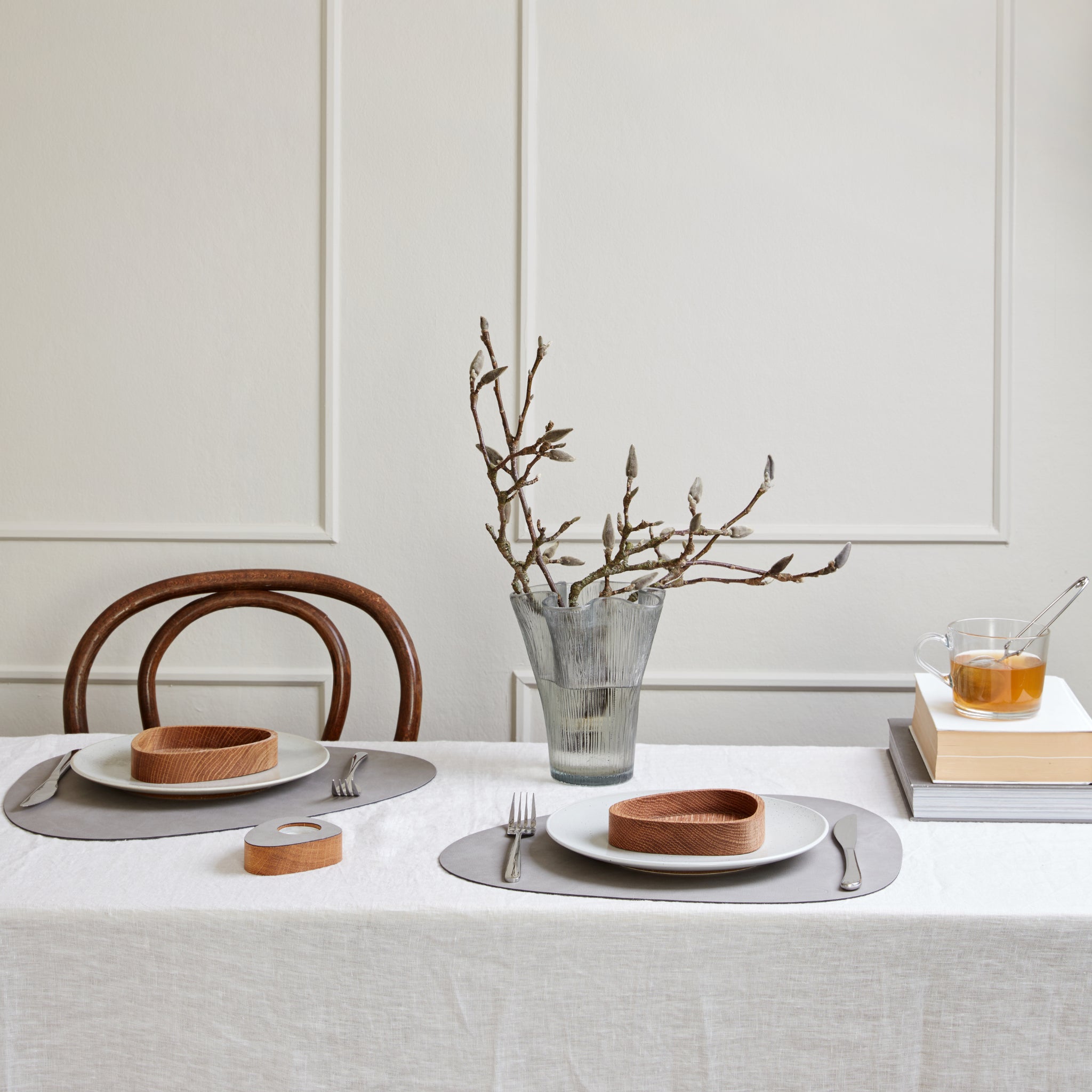 Een minimalistische eetset voor twee personen heeft witte borden, houten kommen, zilver bestek en een wit tafelkleed. De Lind DNA Serene Placemat Curve Ash 37/44 voegt textuur toe naast een glazen vaas en theepot op gestapelde boeken tegen een lichte wand met panelen.