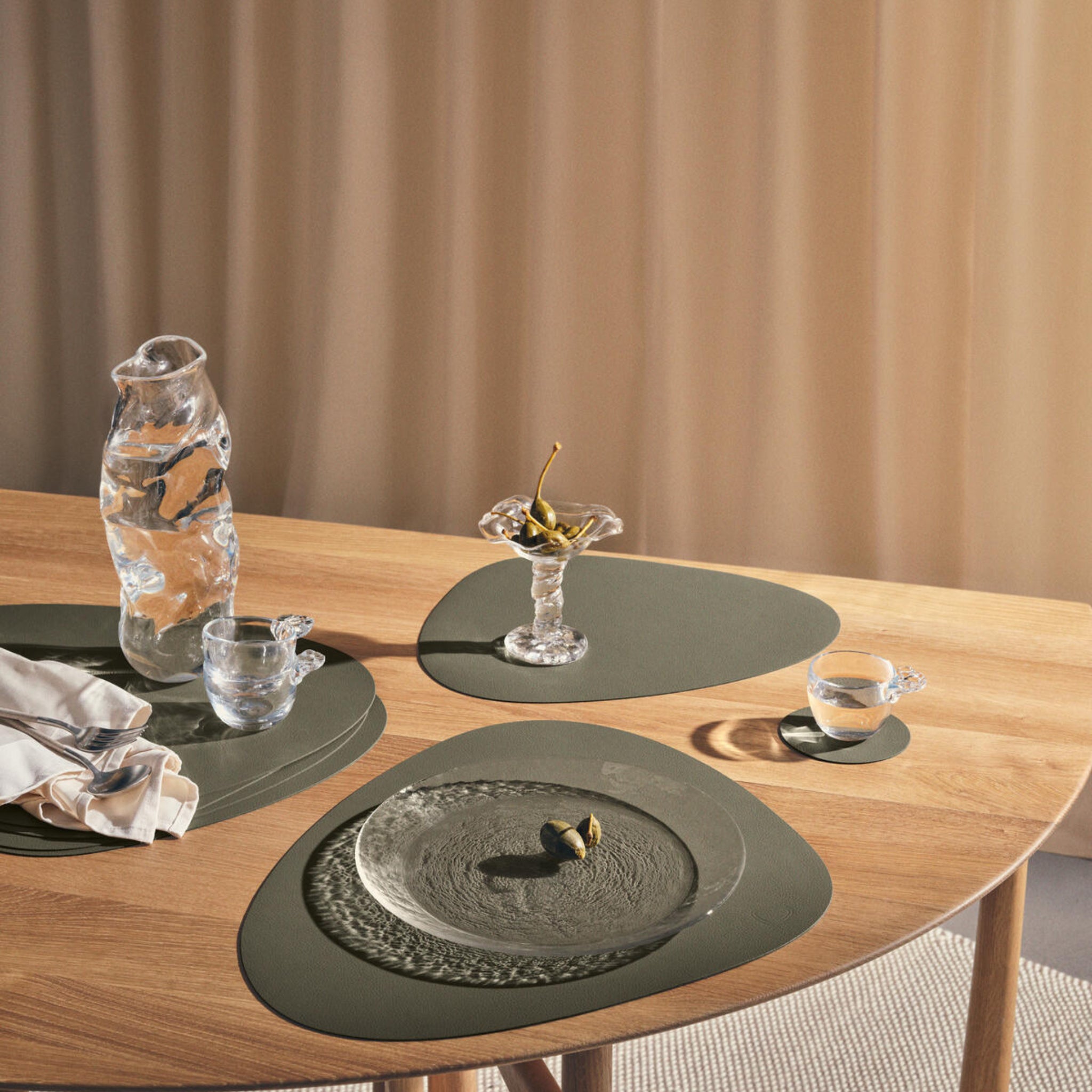 Een houten eettafel gedekt met gestructureerd glaswerk, een karaf, twee kopjes, een bord olijven, een dessertschaal met druiven, een servet en de moderne Lind DNA Serene Placemat Curve Mos 37/44 in groen gerecycled leer. Op de achtergrond hangt een beige gordijn.