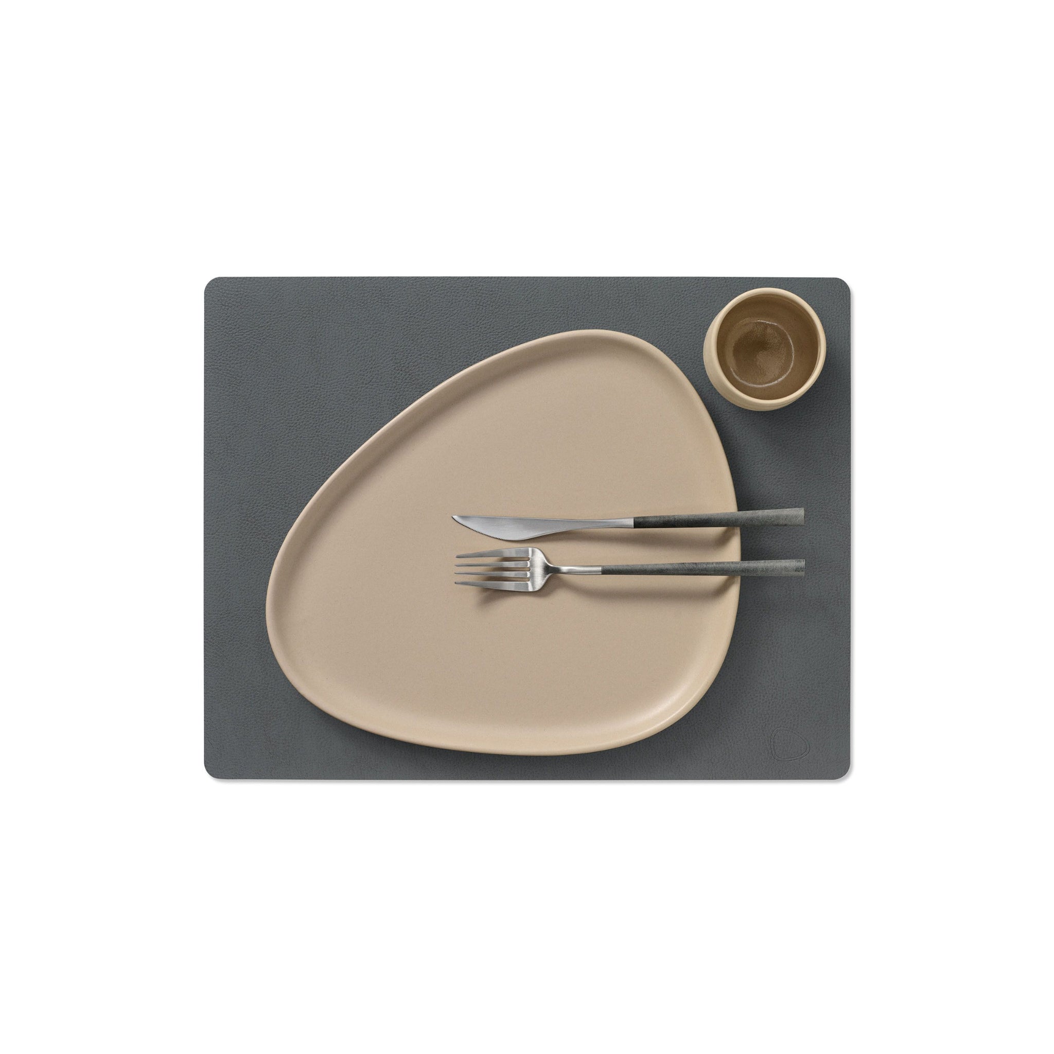 Een beige, asymmetrisch bord met bestek en een bijpassende beker liggen op de Lind DNA Serene Placemat Rechthoekig Antraciet 35/45, met een gerecycled leren textuur van Lind DNA, tegen een witte achtergrond.