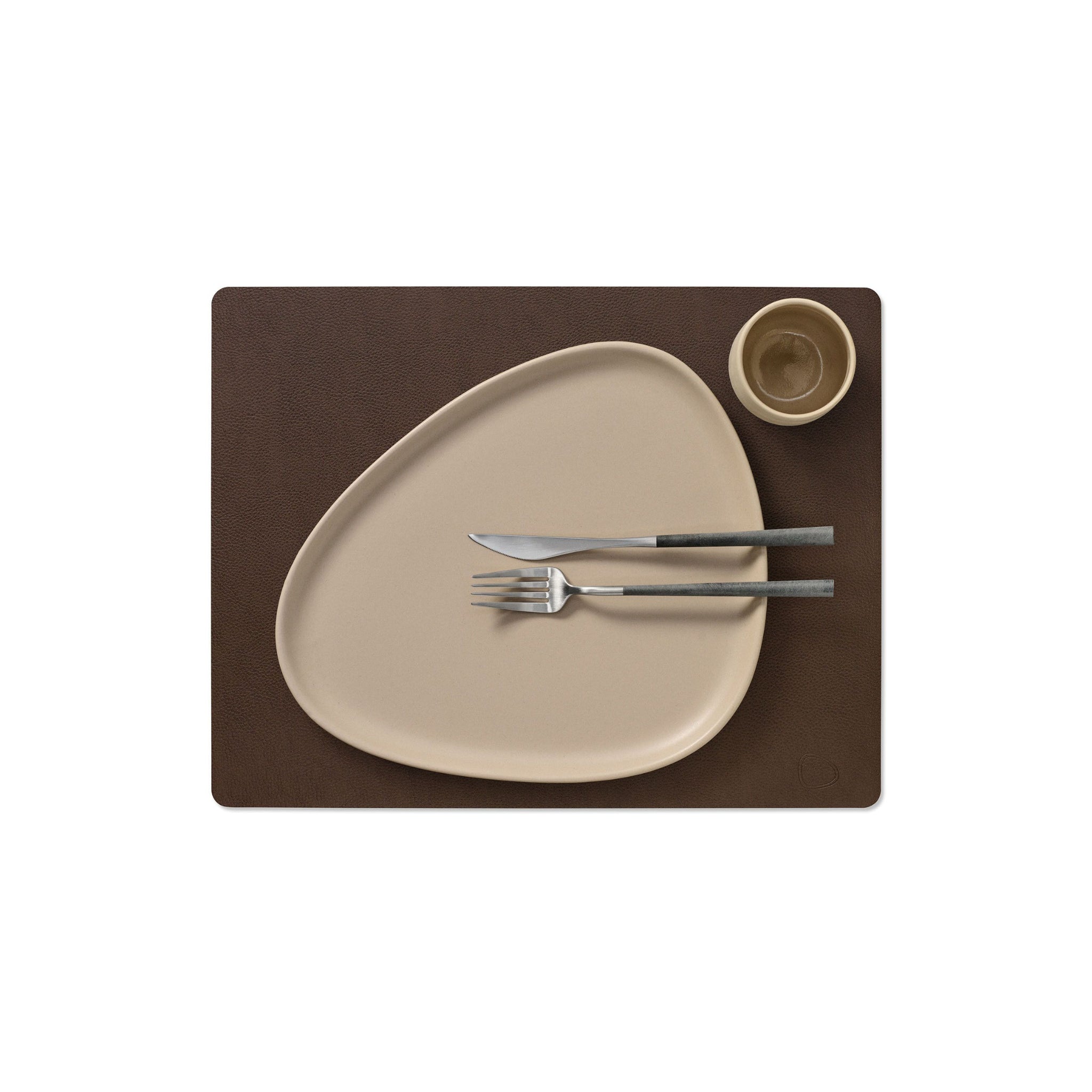 Een Lind DNA Serene Placemat Rechthoekig Hazel 35/45 bevat een beige asymmetrisch bord, bijpassende beker en bestek met metalen handvat in een nette, minimalistische opstelling.
