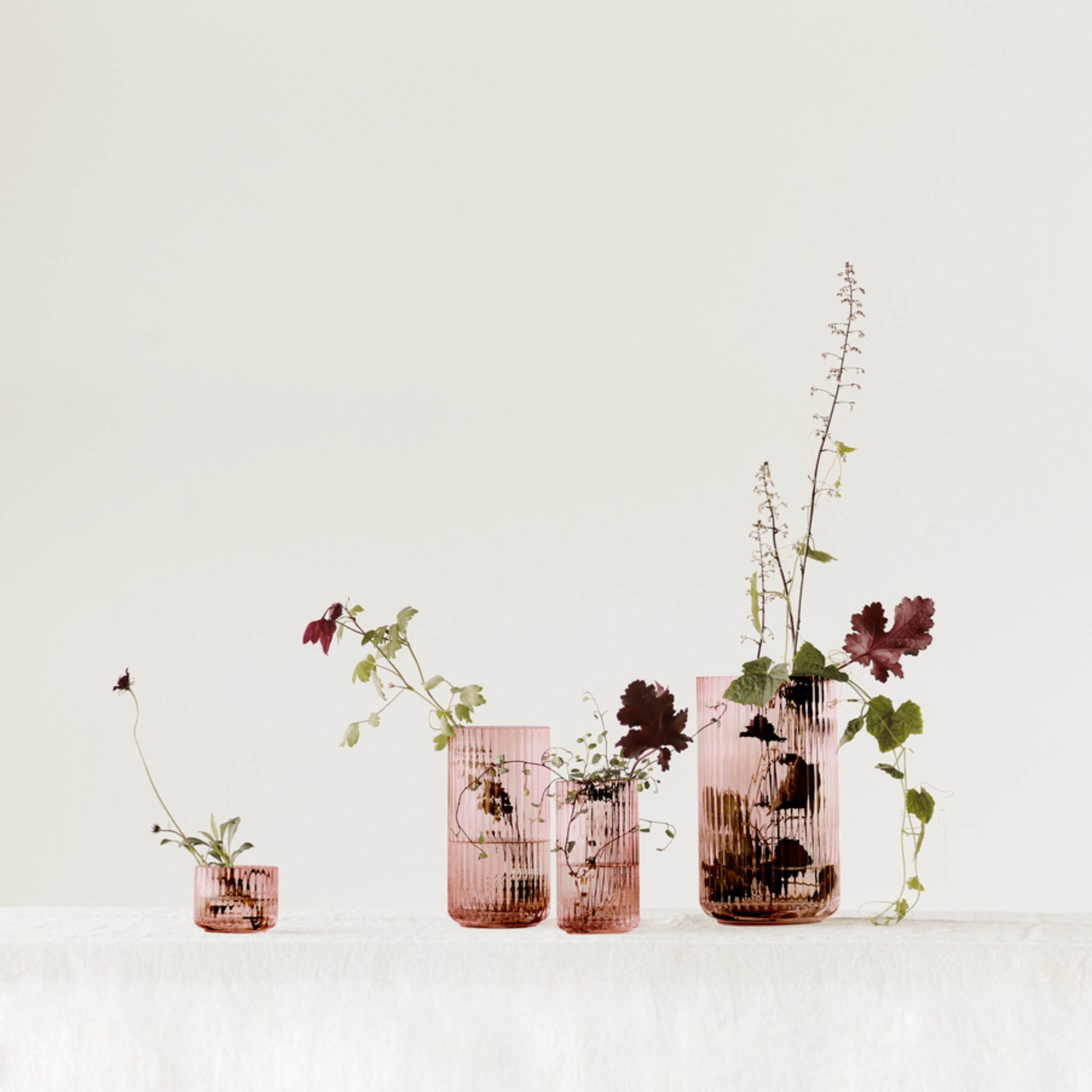 Vier Lyngby Porcelaen Vaas Bordeaux H20 vazen, elk gevuld met groene en donkerrode bladplanten, staan op een rij op een wit oppervlak tegen een eenvoudige, lichte achtergrond.