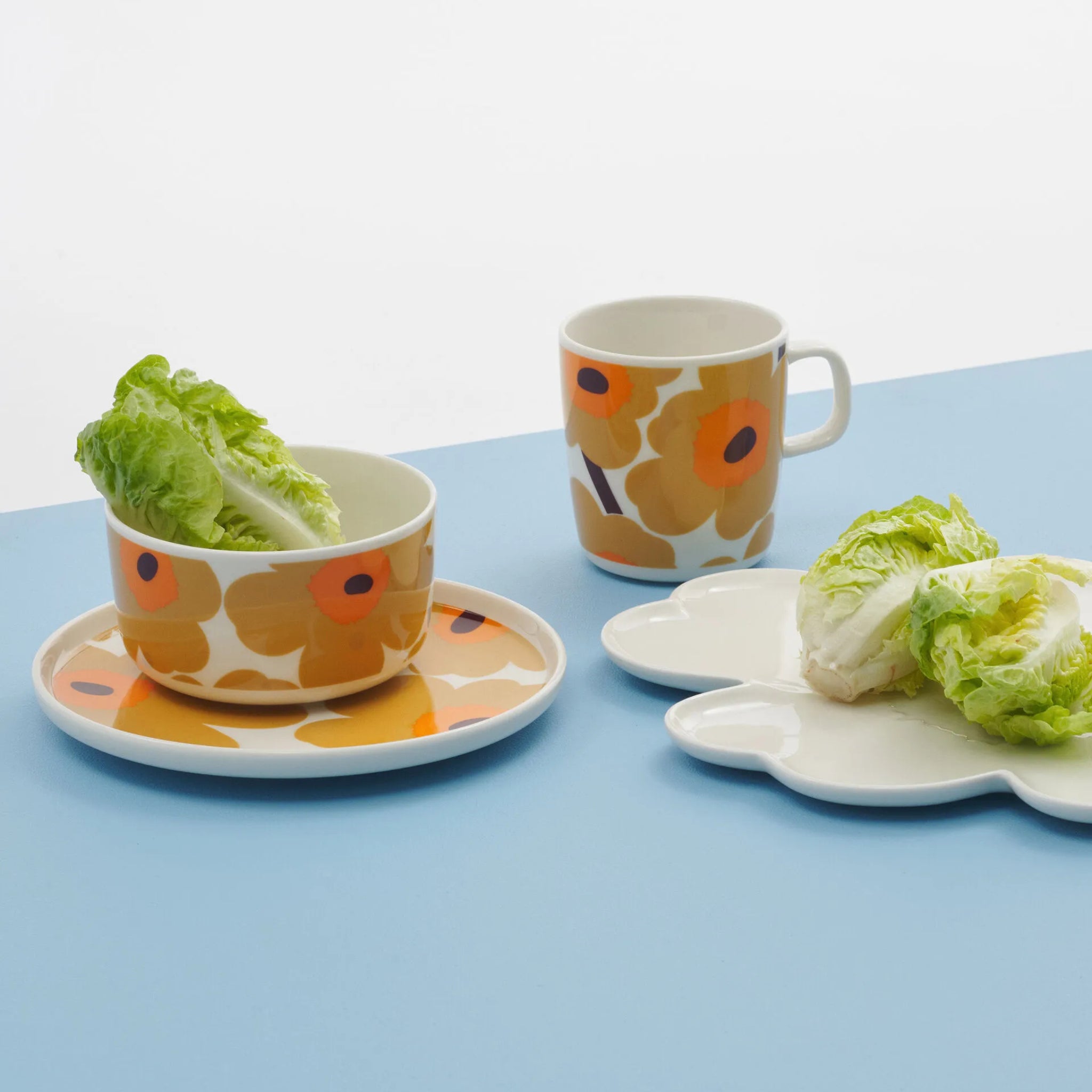 Marimekko's Oiva Unikko Kom Geel D12,5, een kom van keramiek met een opvallende oranje en gele bloemenprint, plus een bijpassende beker en bord, zijn uitgestald met Romeinse sla. Een wit bord met gebogen randen bevat ook sla op een lichtblauw oppervlak.