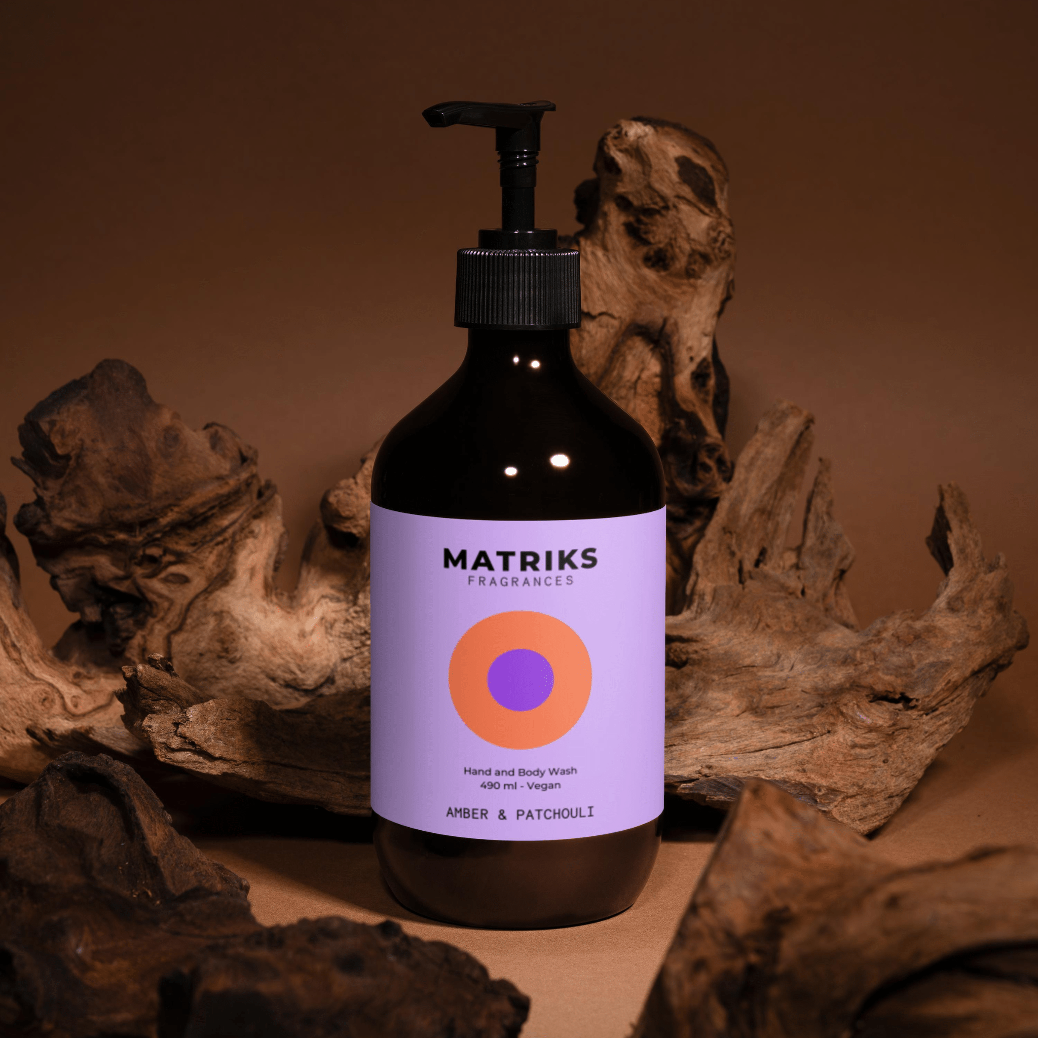 Een 490 ml fles Matriks Fragrances Hand & Body Wash Amber & Patchouli, met een paars etiket met een oranje-roze cirkel, staat tussen drijfhout op een bruine achtergrond.