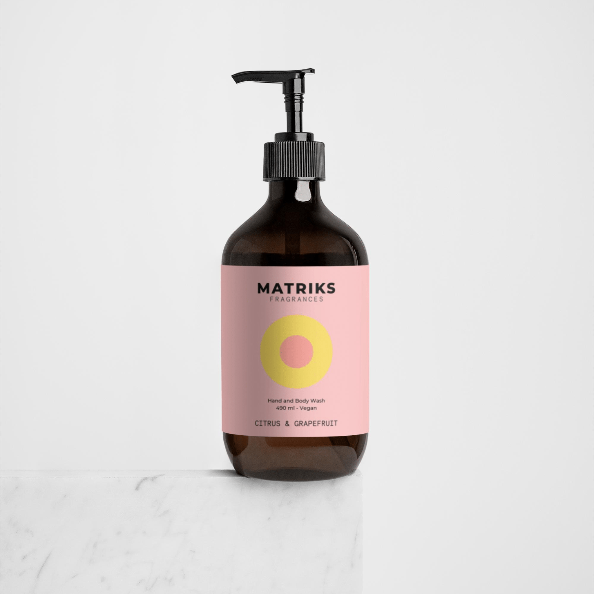 Een bruine pompflacon van Matriks Fragrances Hand & Body Wash Citrus & Grapefruit 490 ml met een roze label en gele cirkel staat op wit marmer, met een veganistische formule en verfrissende grapefruitgeur tegen een effen witte achtergrond.