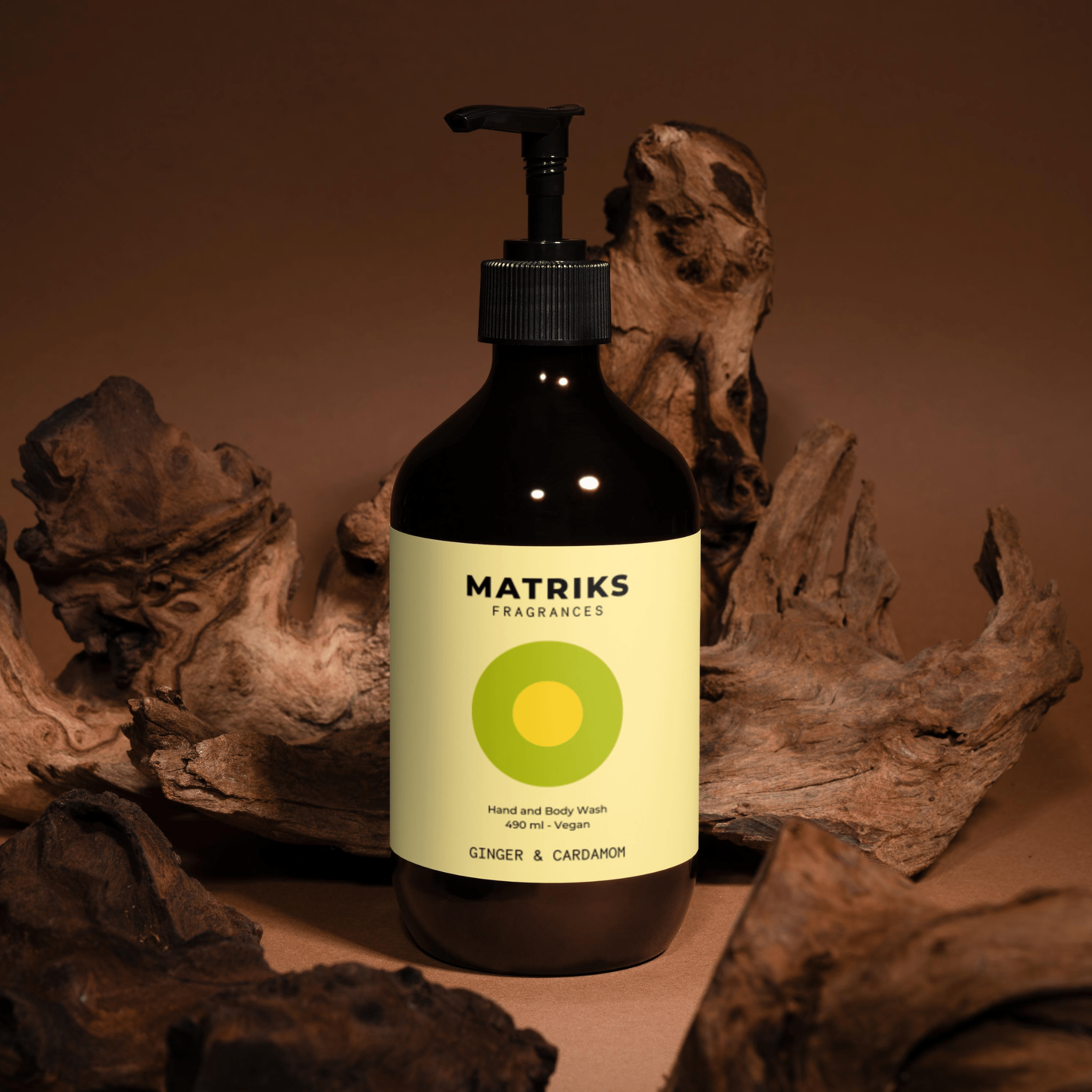 Een bruine fles Matriks Fragrances Hand & Body Wash Ginger & Cardamom 490 ml rust tussen het drijfhout, met een geelgroen etiket - en brengt unieke geuren naar je handzeeproutine met zijn veganistische gember- en kardemomgeur.