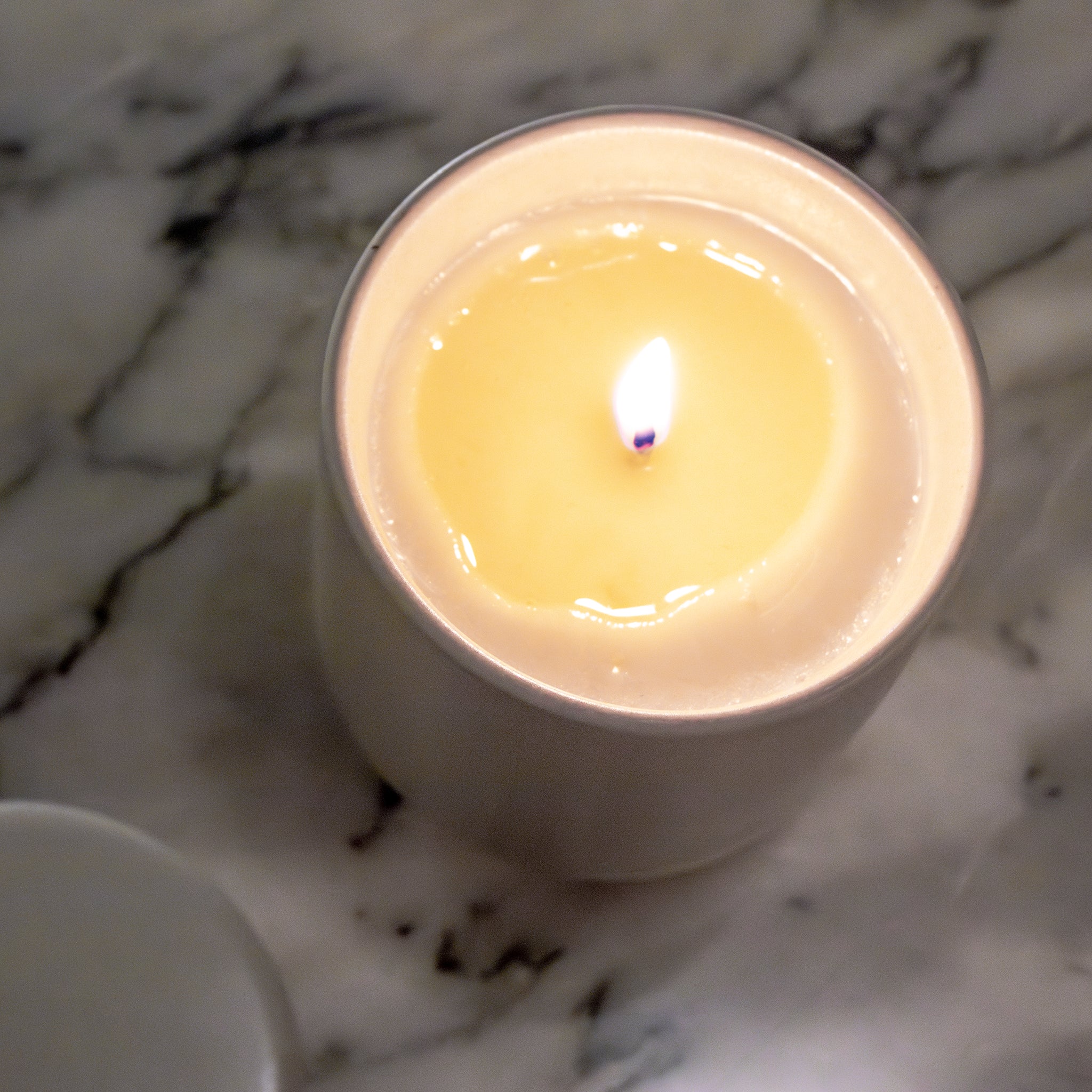 Een Matriks Fragrances Geurkaars Keramiek Eikenhout Embers 270 g in een wit potje rust op marmer, het deksel ernaast. De heldere vlam werpt een warme gloed op de romige was die in Antwerpen is handgemaakt voor authentieke lokale charme.