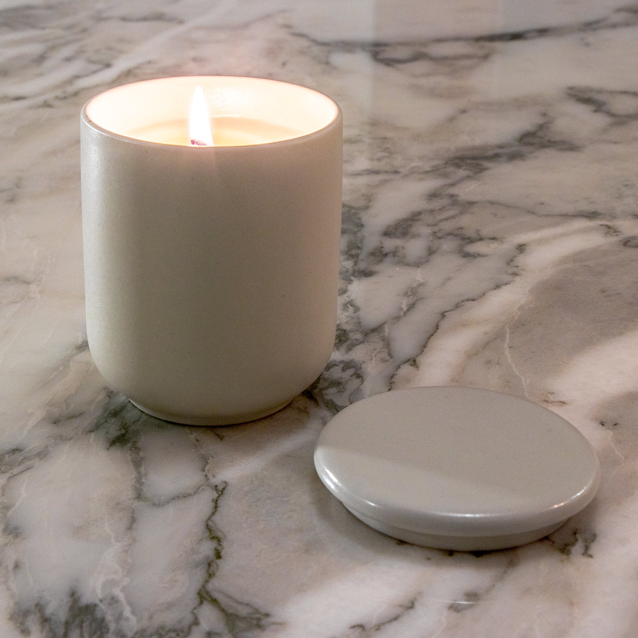 Een lit Matriks Fragrances Geurkaars Keramiek Sweet & Smokey 270 g van Matriks Fragrances staat op marmer, handgemaakt in Antwerpen, met het ronde deksel ernaast.