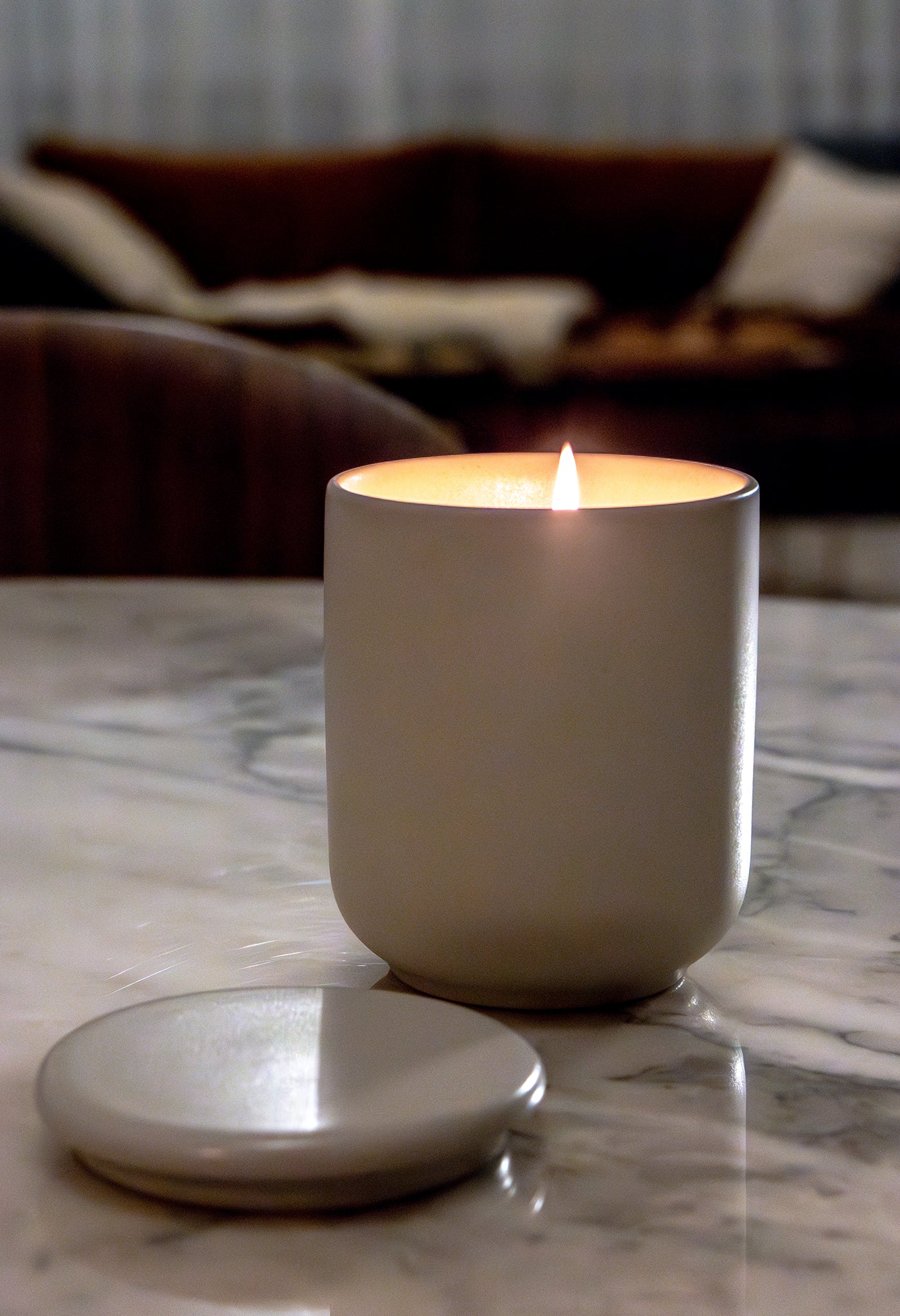 De Matriks Fragrances Geurkaars Keramiek Cabin Glow 270 g zit in een eenvoudige witte pot op een marmeren tafel, met het deksel vlakbij. Een vage bank met kussens op de achtergrond versterkt de gezellige sfeer. Handgemaakt in Antwerpen.