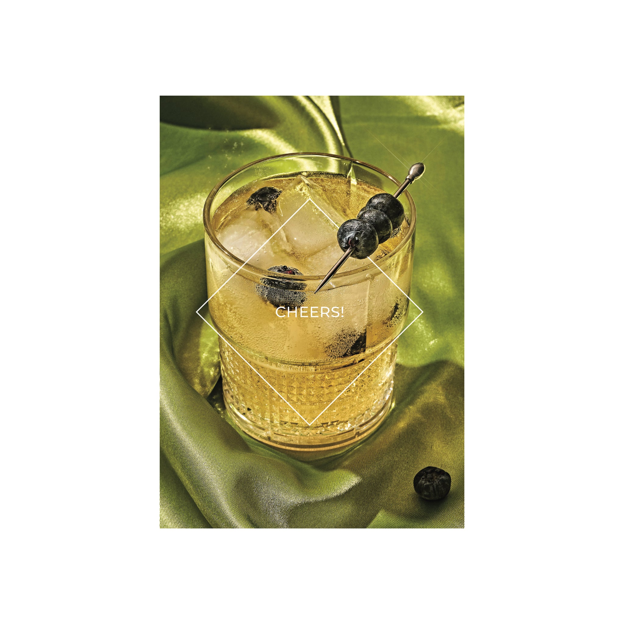 Een glas gouden ijscocktail met bosbessen op een stokje rust op glanzend groene stof onder "CHEERS!" in een witte ruit. Perfect voor de stijlvolle Matriks Postkaart Lijn Cheers Dubbel A6 van Matriks, geïnspireerd op Antwerpse gebouwen.