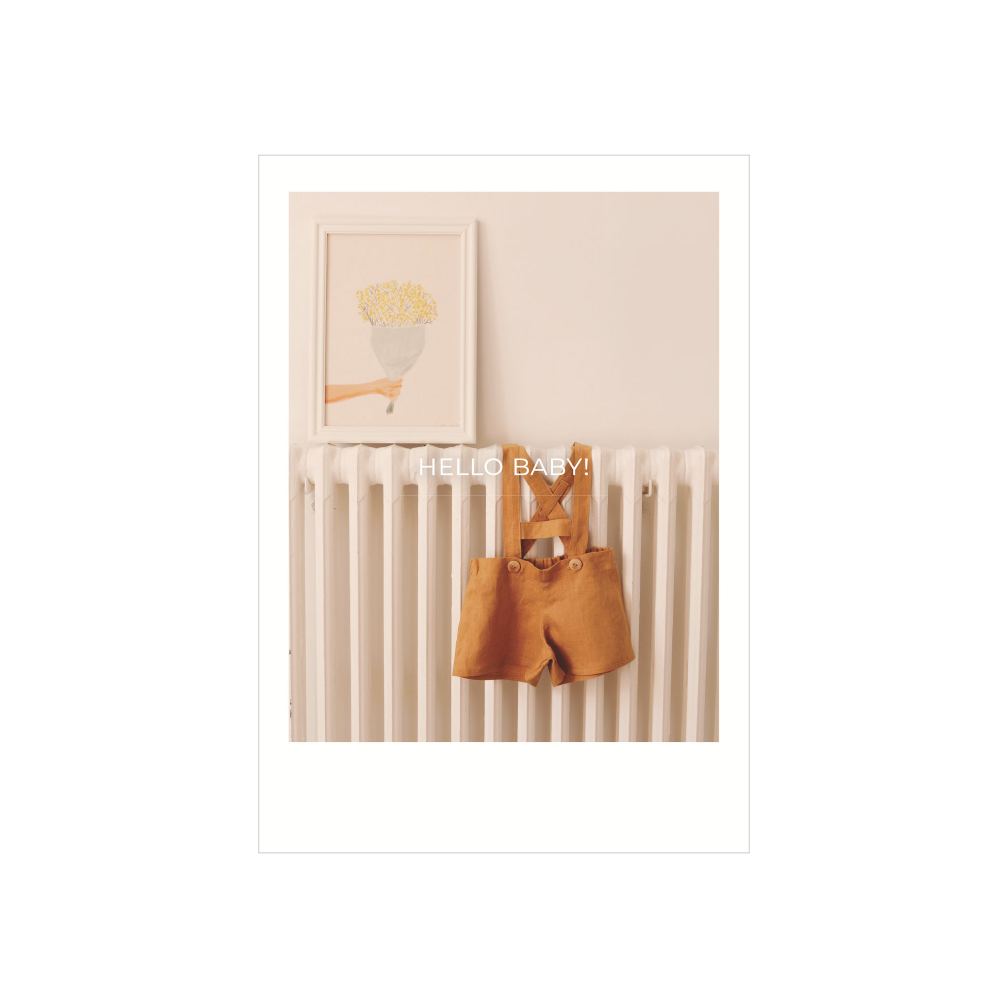 Een paar kleine mosterdkleurige babyoveralletjes hangt aan een witte radiator met bloemen erboven en "HELLO BABY!" tekst - ideaal voor unieke Matriks Postkaart Polaroid Hello Baby Dubbel A6 wenskaarten.