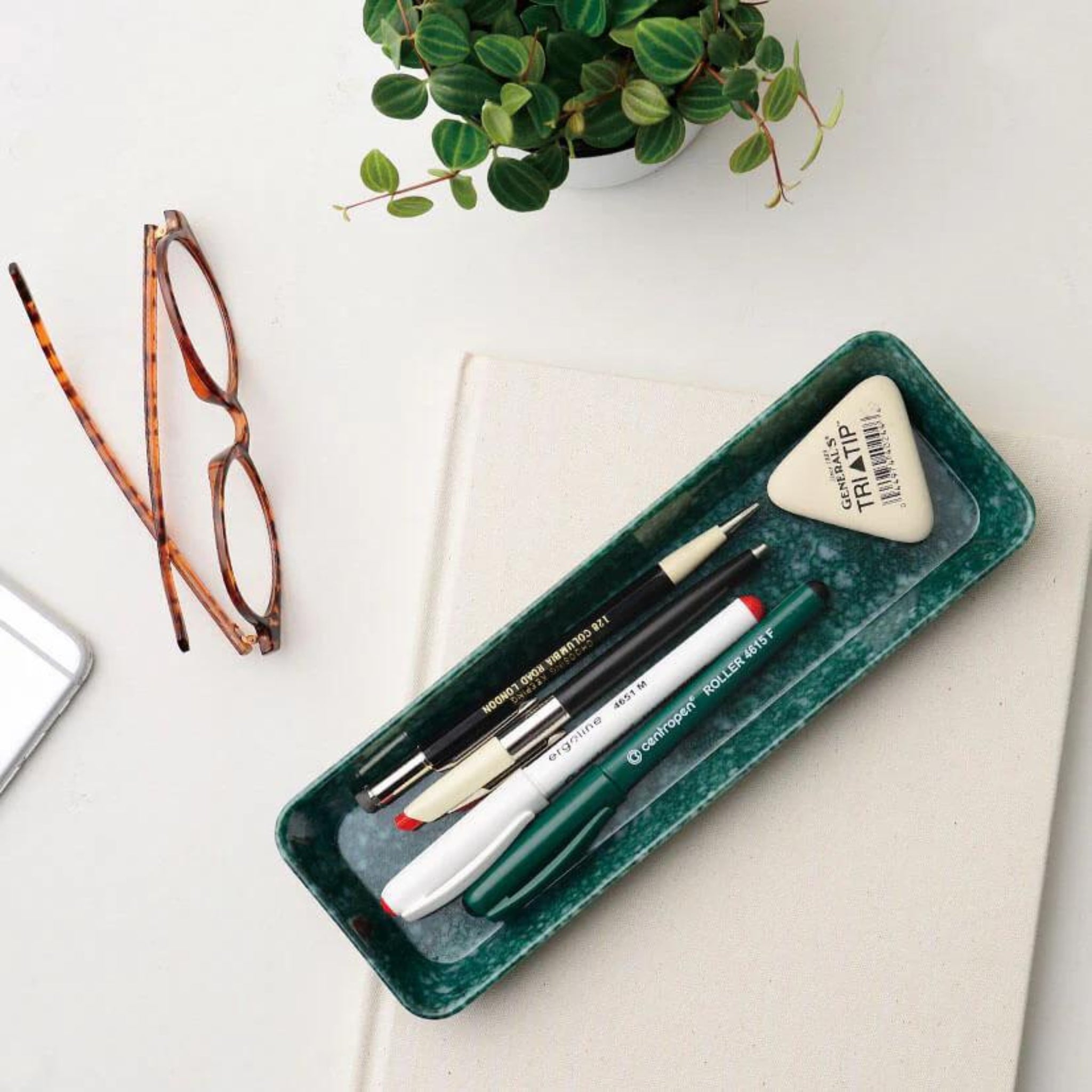 In de Melamine Marbled Bakje Donkergroen 23/7 van Melamine Marbled passen drie pennen, een vulpotlood en een driehoekige gum. Vlakbij staan een bril met schildpad, een witte potplant en een gesloten laptop - ideaal voor het organiseren van je bureau.