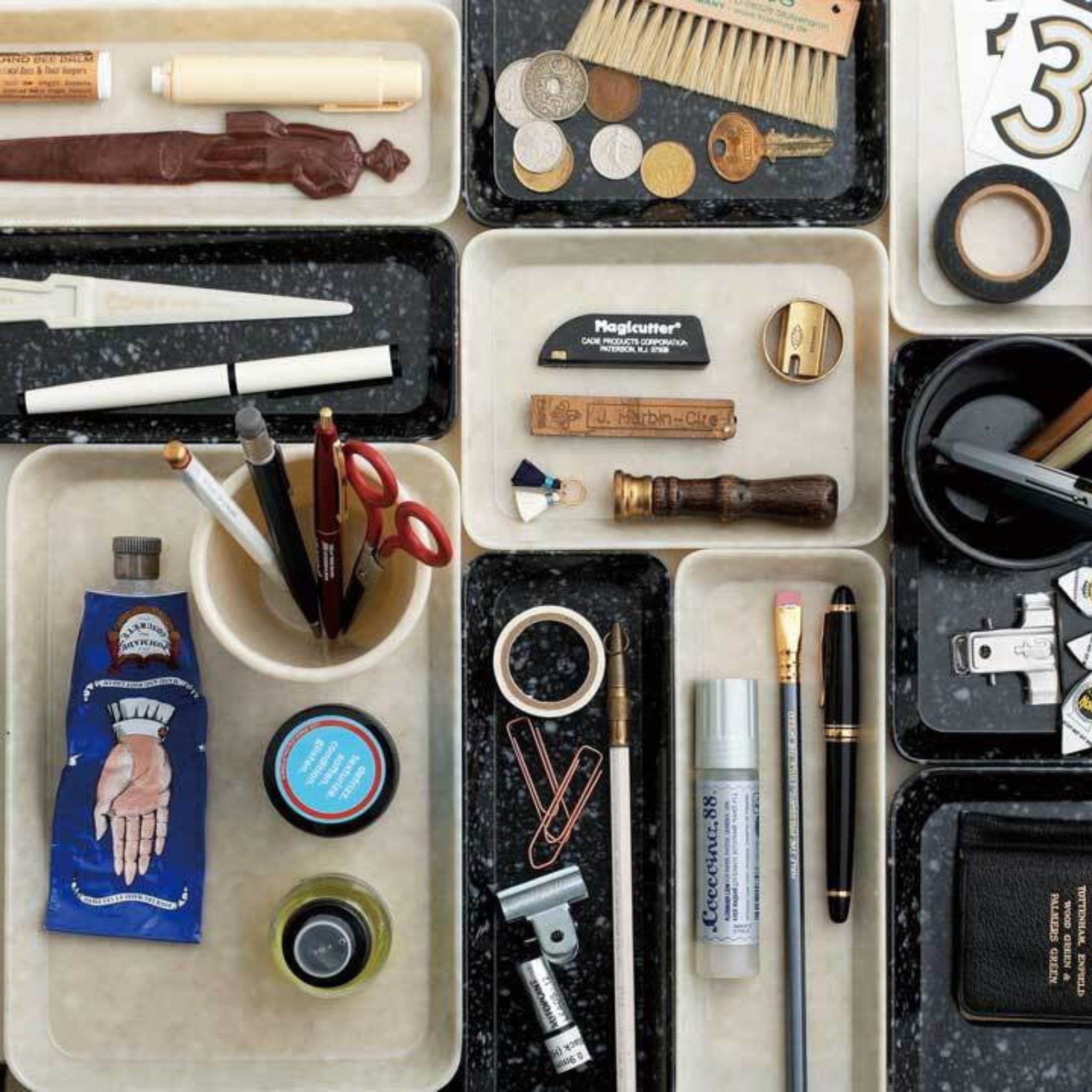 Een selectie van kantoorbenodigdheden zoals scharen, pennen, munten en meer in het Melamine Marbled Bakje Ivoor 23/7 van Melamine Marbled, gerangschikt op een gespikkeld zwart oppervlak.