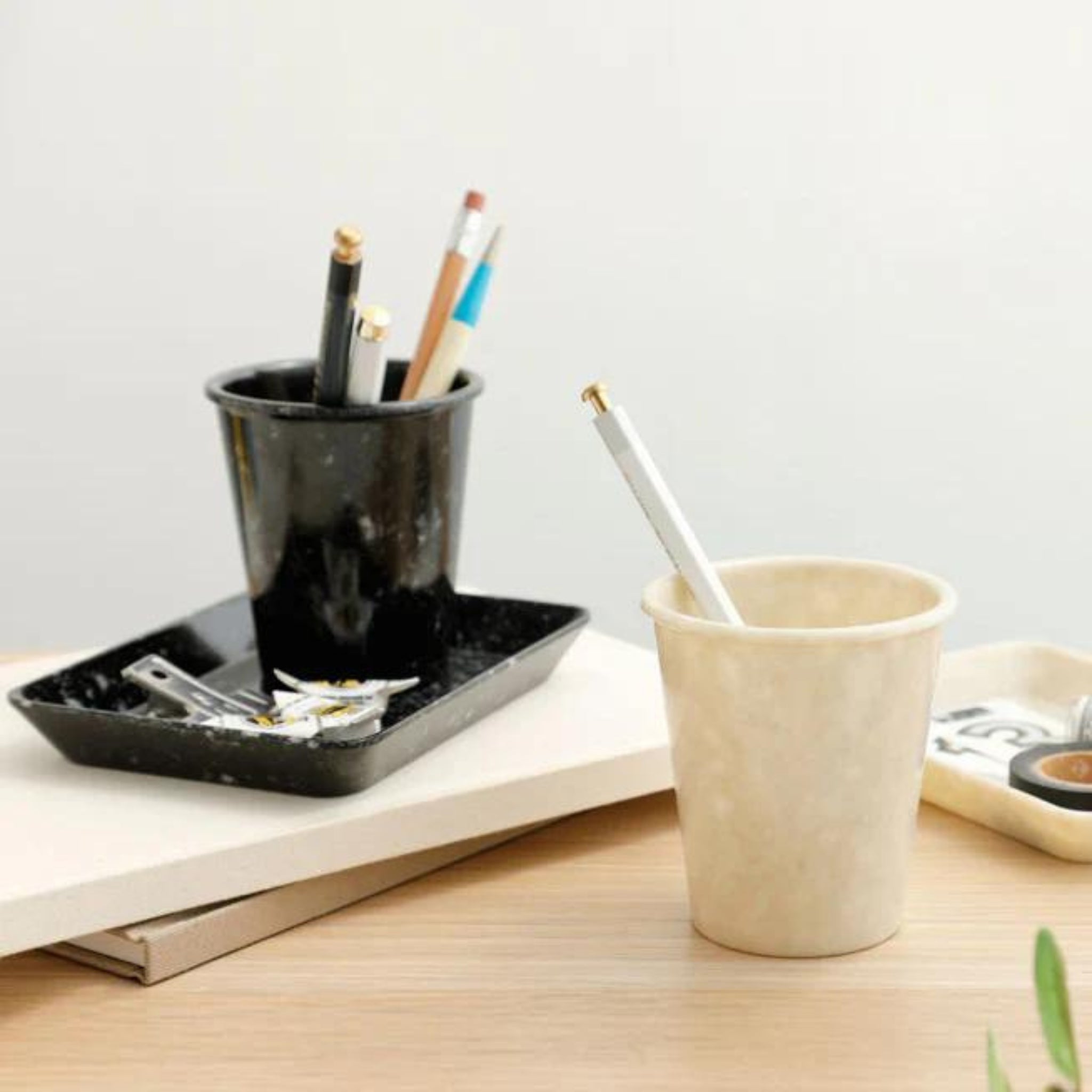 Een gemarmerde Melamine Beker Zwart en een beige beker, beide gevuld met pennen en potloden, rusten op een houten bureau naast een organizer tray met paperclips en scharen, tegen een minimalistische neutrale achtergrond.