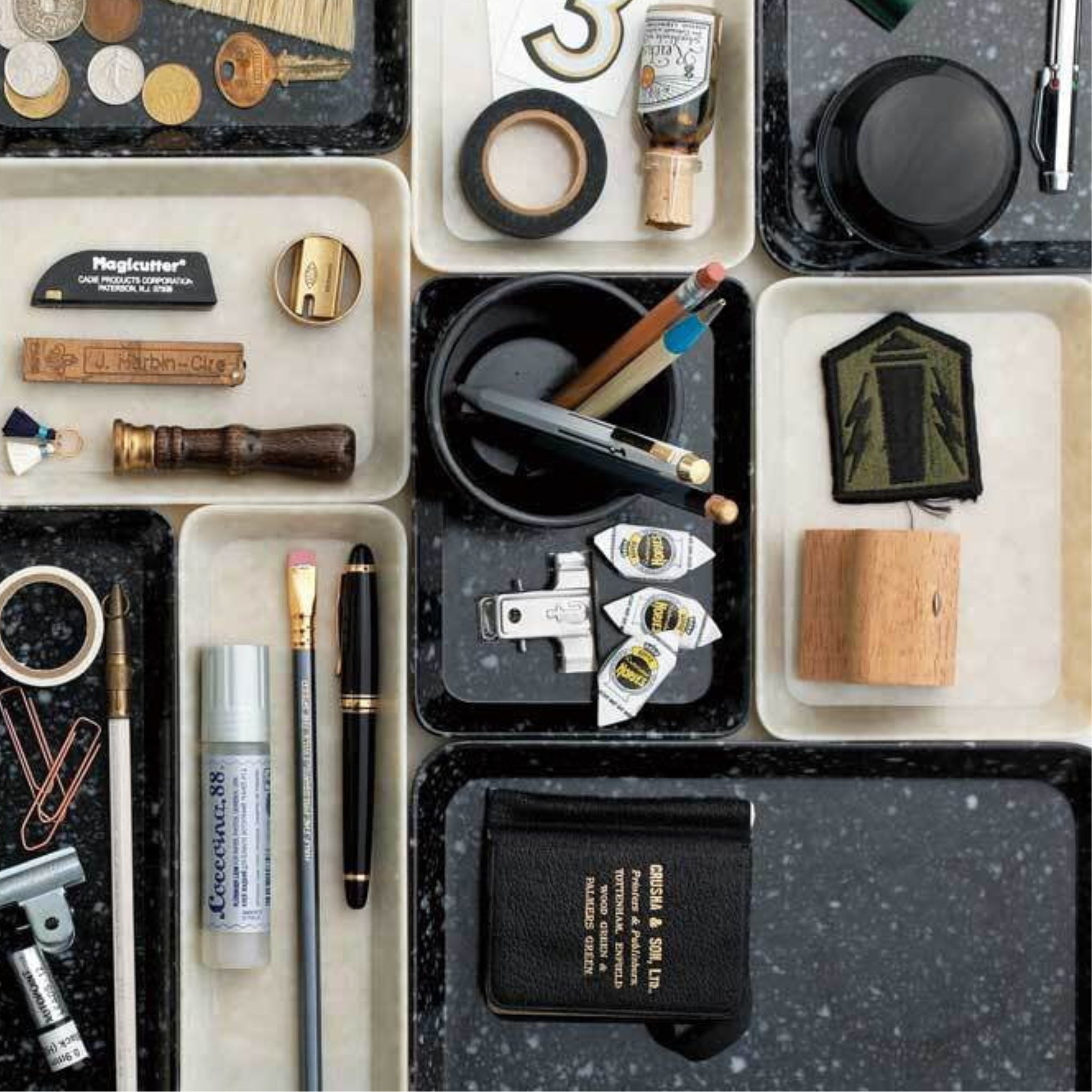 De Melamine Marbled Beker Zwart van Melamine Marbled staat op een gespikkeld oppervlak, netjes gerangschikt tussen kantoorbenodigdheden zoals pennen, potloden, organizers, puntenslijpers, clips, stempels en kleine bakjes in verdeelde bakjes.