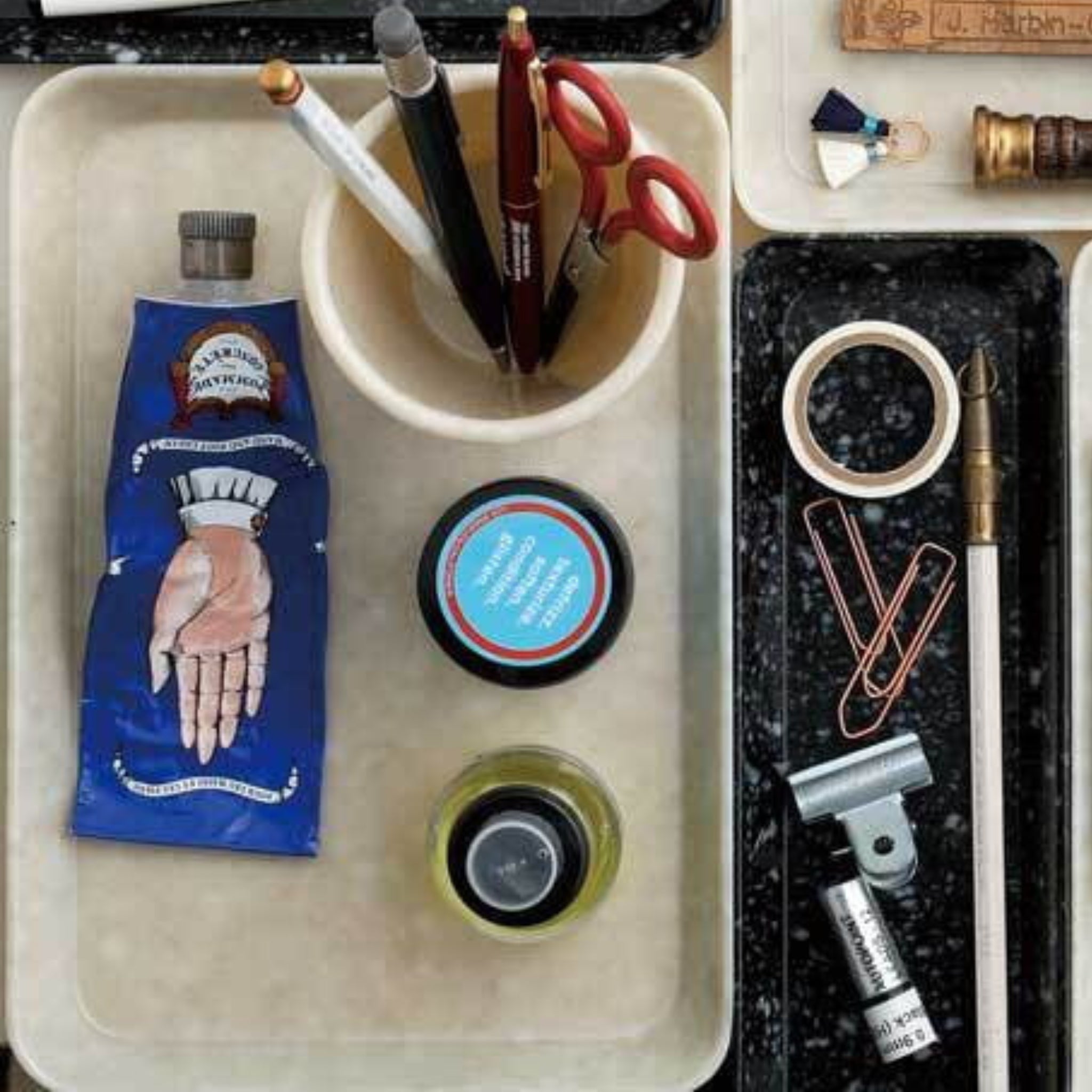 Een gemarmerde Dienblad Ivoor 23/16 van Melamine Marbled laat kunstbenodigdheden netjes zien: een blauwe verftube, inktflesjes, pennen en scharen in een beker, plakbandrol, paperclips, ordnerclip en een kalligrafiepen.