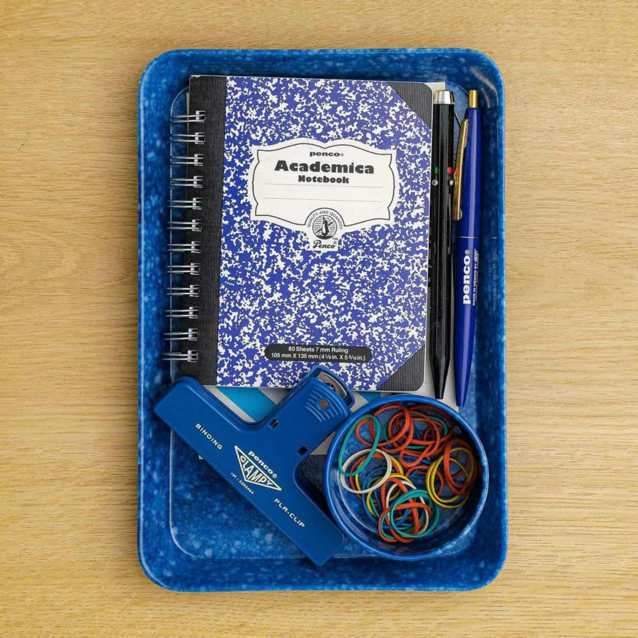 De Melamine Marbled Dienblad Navy 23/16 van Melamine Marbled ligt op een houten oppervlak en biedt ruimte aan een spiraalvormig notitieboek, twee pennen, drie potloden, een blauwe nietmachine en een ronde houder met kleurrijke elastiekjes - een ideale kantoorhouder.