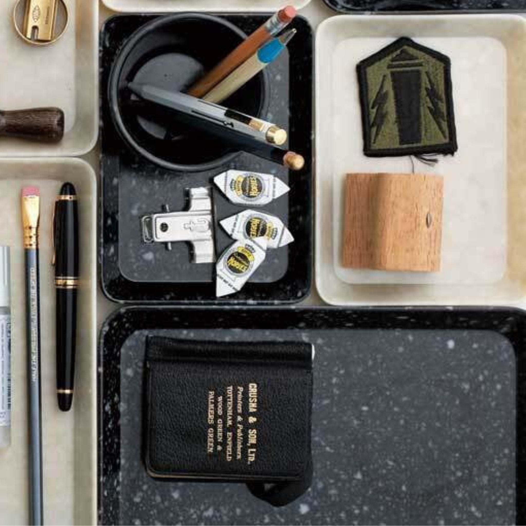 Een kantoorhouder op een Melamine Marbled Dienblad Zwart 23/16 van Melamine Marbled bevat pennen, potloden, een vulpen, paperclips, puntenslijpers, een houten blok, een patch en een klein leren notitieboekje met goudopdruk.