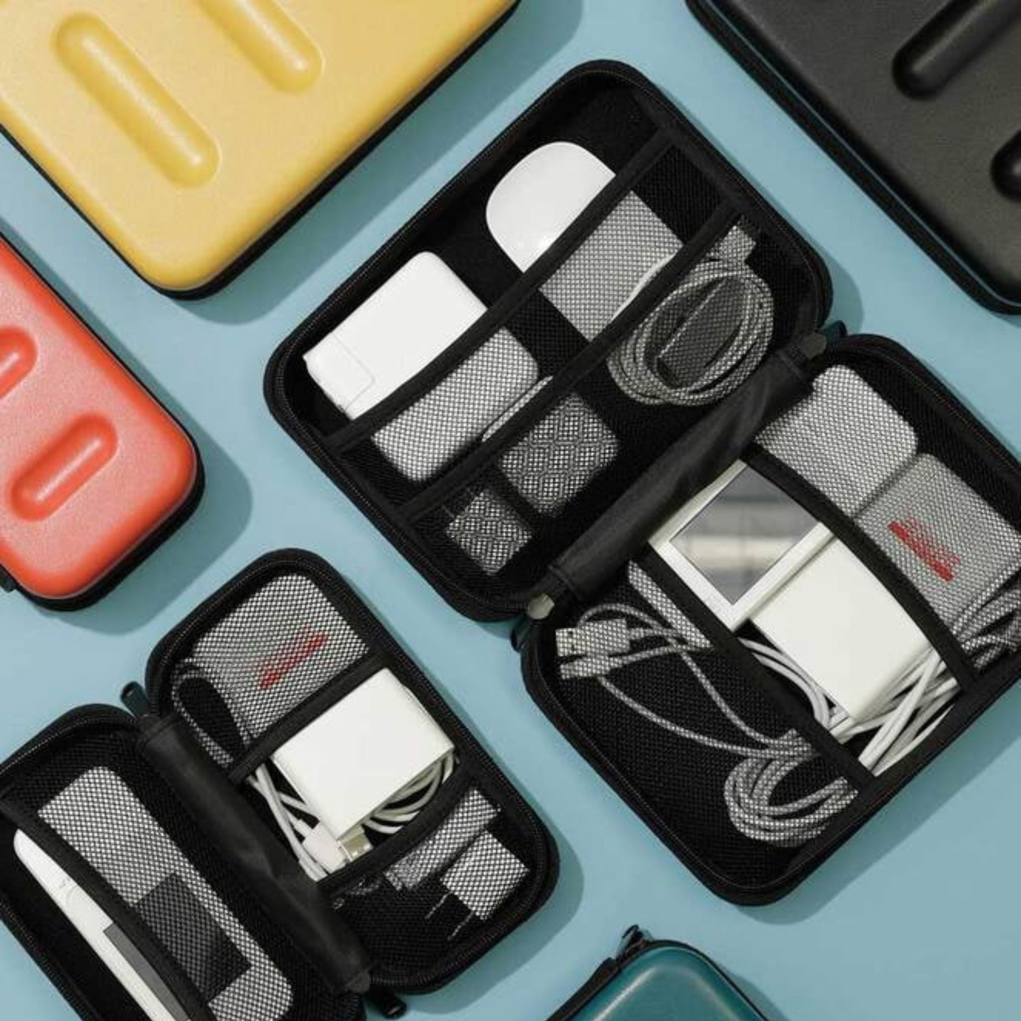 Verschillende Nähe Hard Shell Organizer Ivoor L-koffers van Nähe liggen open op een lichtblauw oppervlak en tonen netjes georganiseerde opladers, kabels en adapters naast een gesloten, kleurrijk polycarbonaat koffertje.