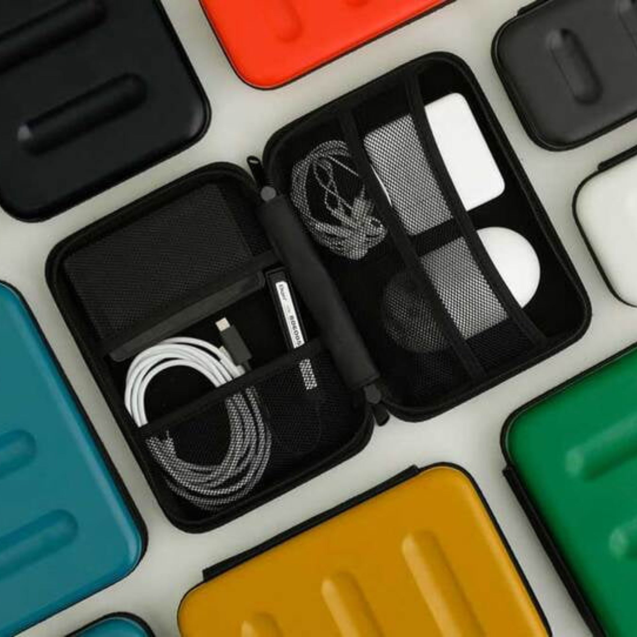 De Nähe Hard Shell Organizer Ivoor L van Nähe, open afgebeeld met vakjes voor een oplaadkabel, adapters en oordopjes, zit tussen gesloten kleurrijke etuis-ideaal voor gadgets opbergen op reis.