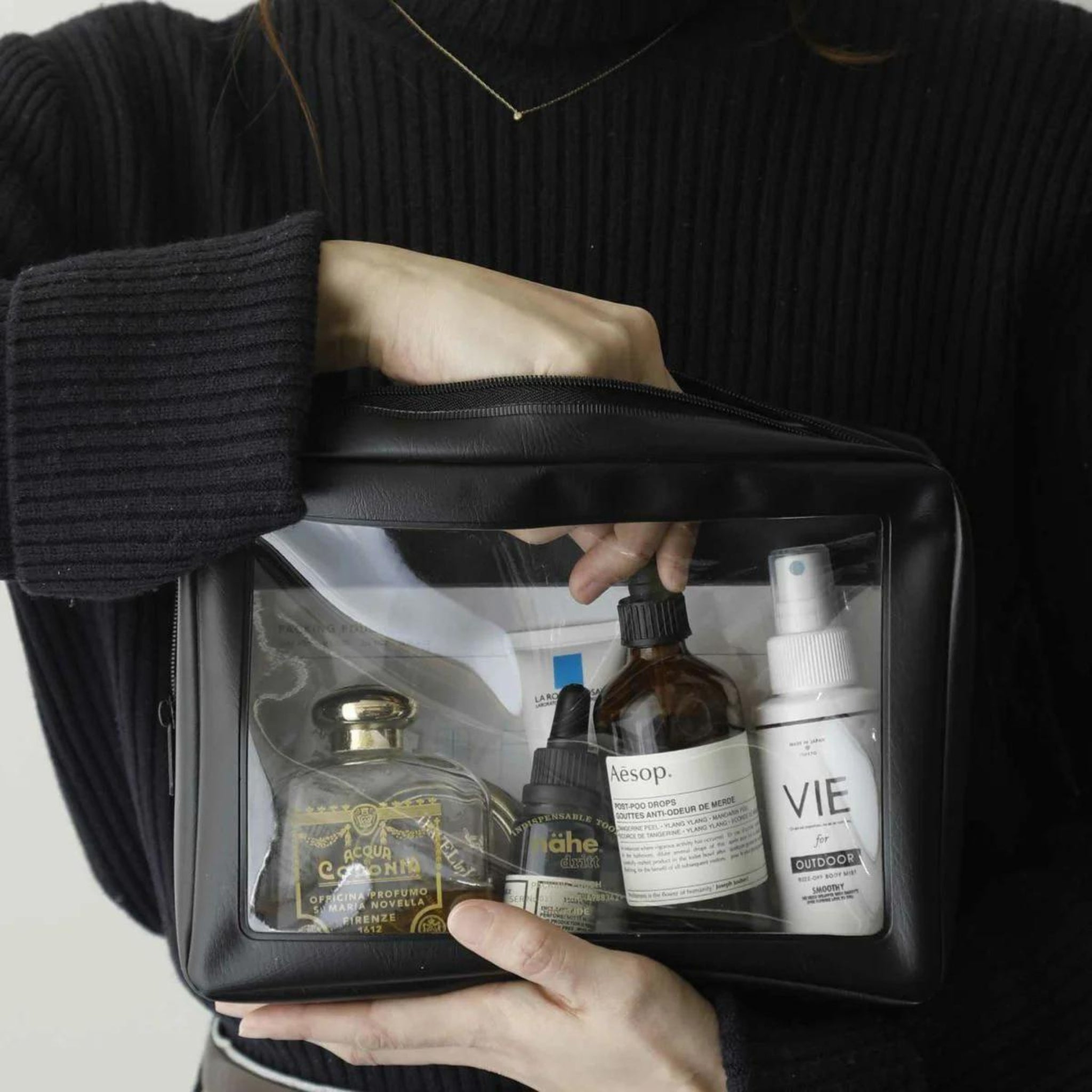 Een persoon in een zwarte trui houdt een Nähe Reiszakje Zwart L van Nähe vast, met daarop gelabelde huidverzorgings- en cosmeticaflessen, waaronder een sprayflesje, die door de transparante voorkant zichtbaar zijn.