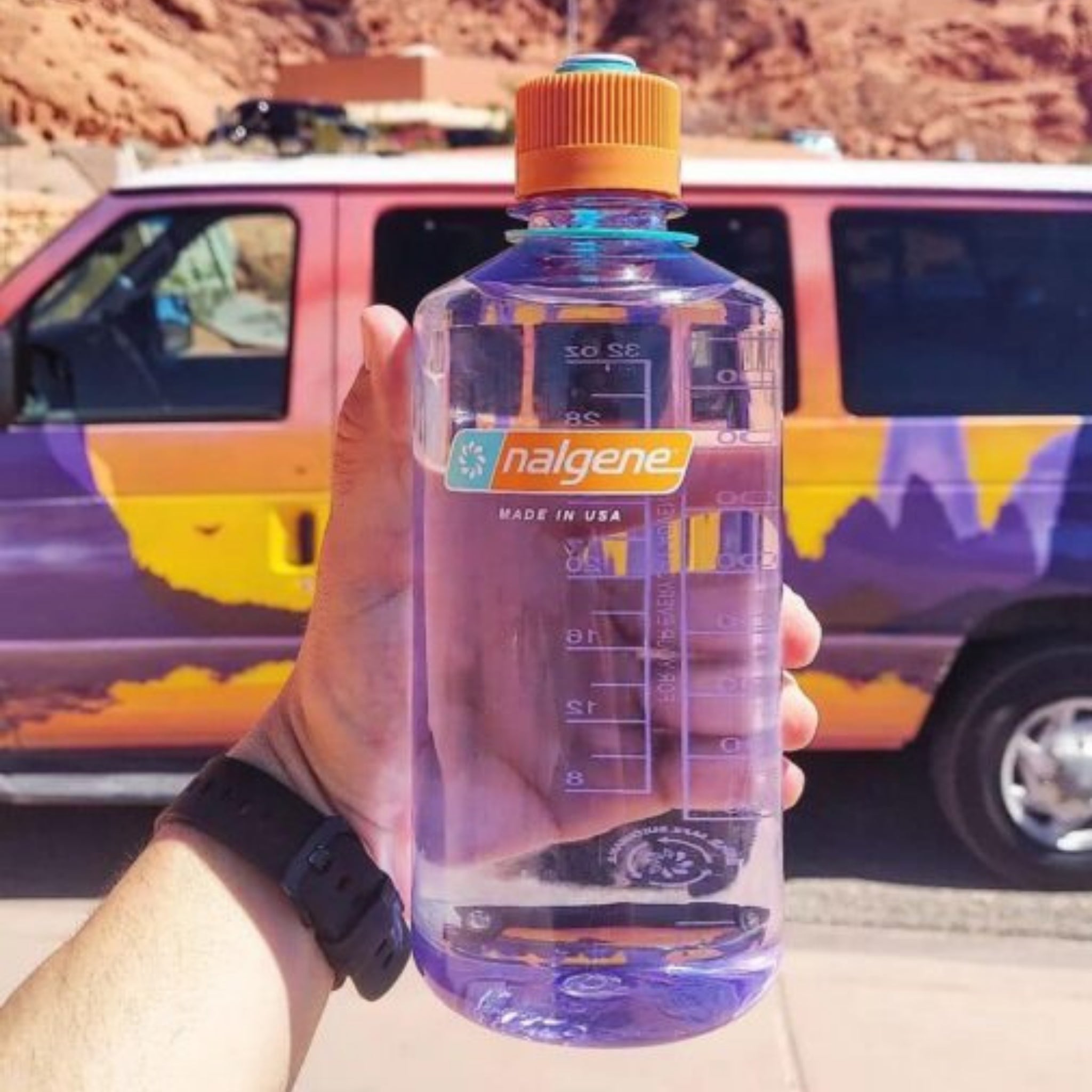 Een hand houdt een Nalgene Drinkfles Smalle Opening Amethyst 1L met oranje dop vast voor een kleurrijk busje en rode rotsheuvels. Deze BPA-vrije fles van Nalgene is ideaal voor elk avontuur.