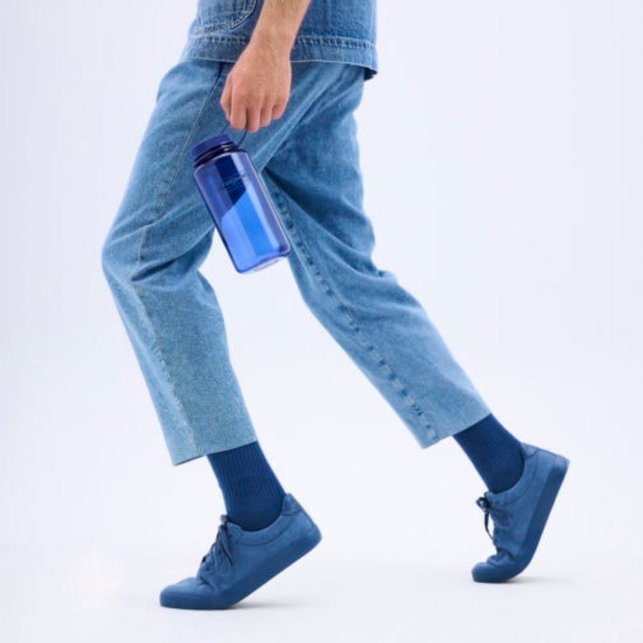 Een persoon in een blauwe spijkerbroek, blauwe sokken en blauwe gympen loopt naar voren met een Nalgene Drinkfles Brede Opening Denim 1L van Nalgene in zijn rechterhand tegen een effen lichte achtergrond.
