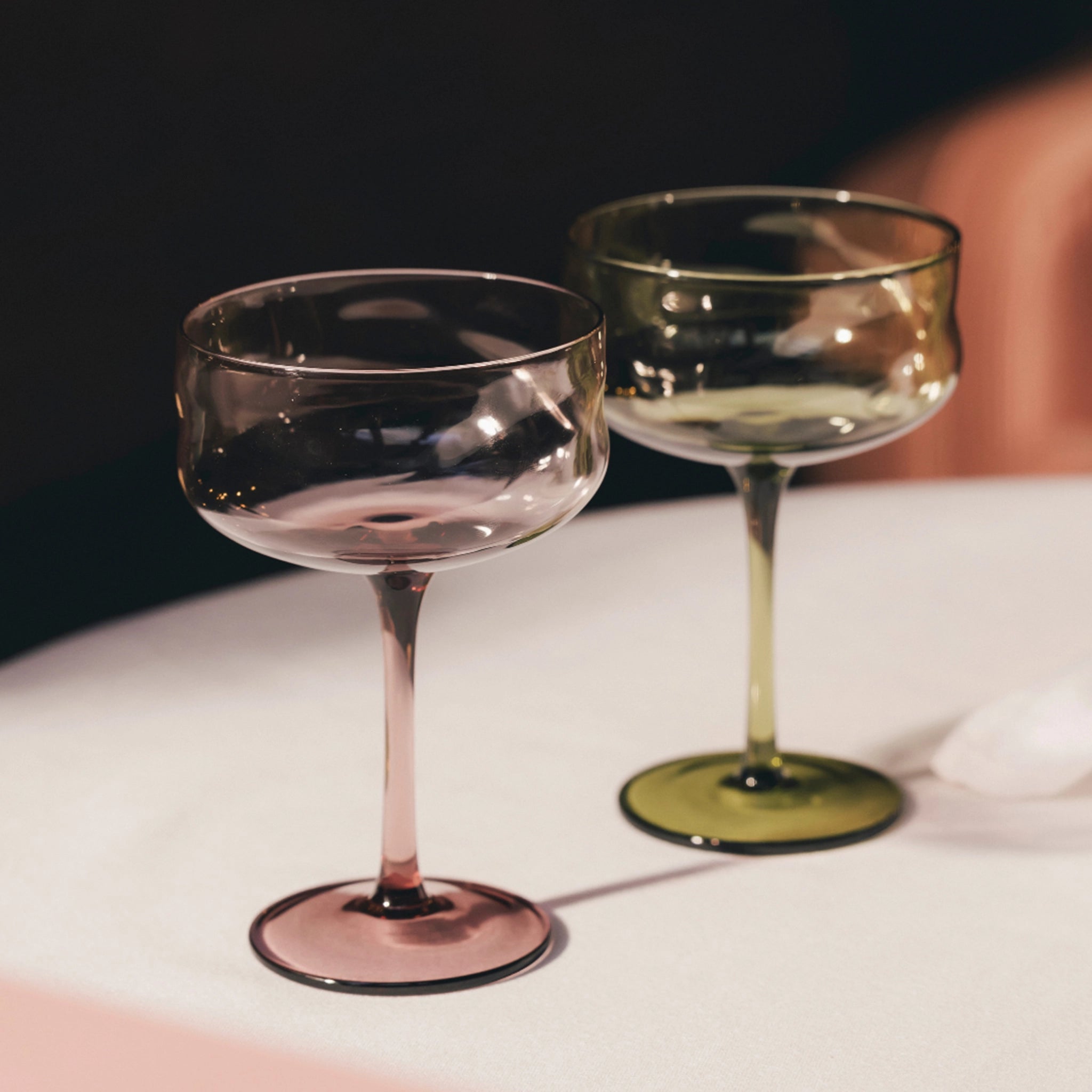 Twee lege Paveau Swirl Champagnecoupes in de kleuren Silt en Bordeaux (26 cl) rusten op een wit tafelkleed onder zachte verlichting en creëren een warme en elegante sfeer - perfect voor feestelijke gelegenheden.