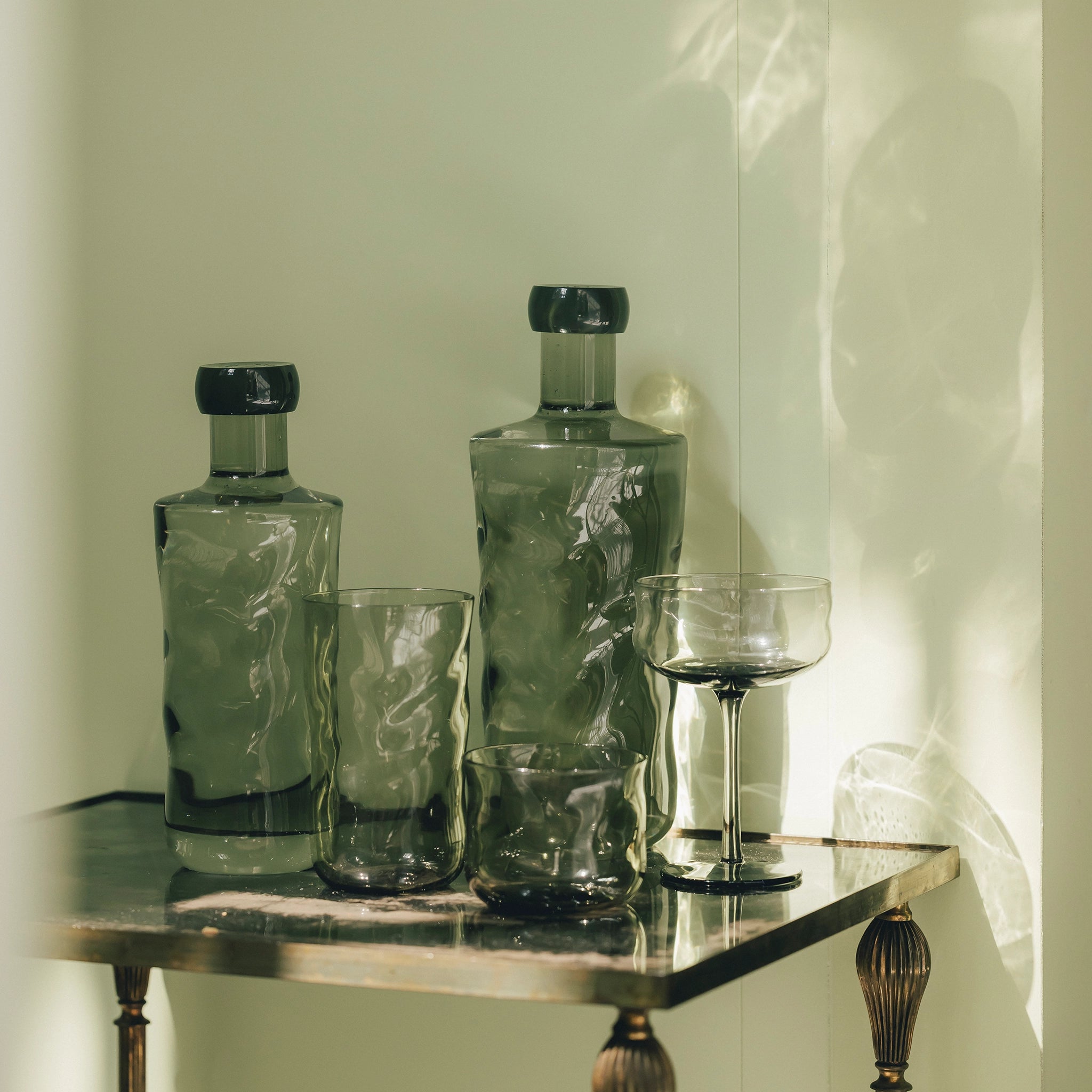 Twee Paveau Swirl Longdrink Glas Mist Smoke 46 cl glazen staan op een metalen tafeltje tegen een bleke muur, het zonlicht werpt rokerige reflecties uit.