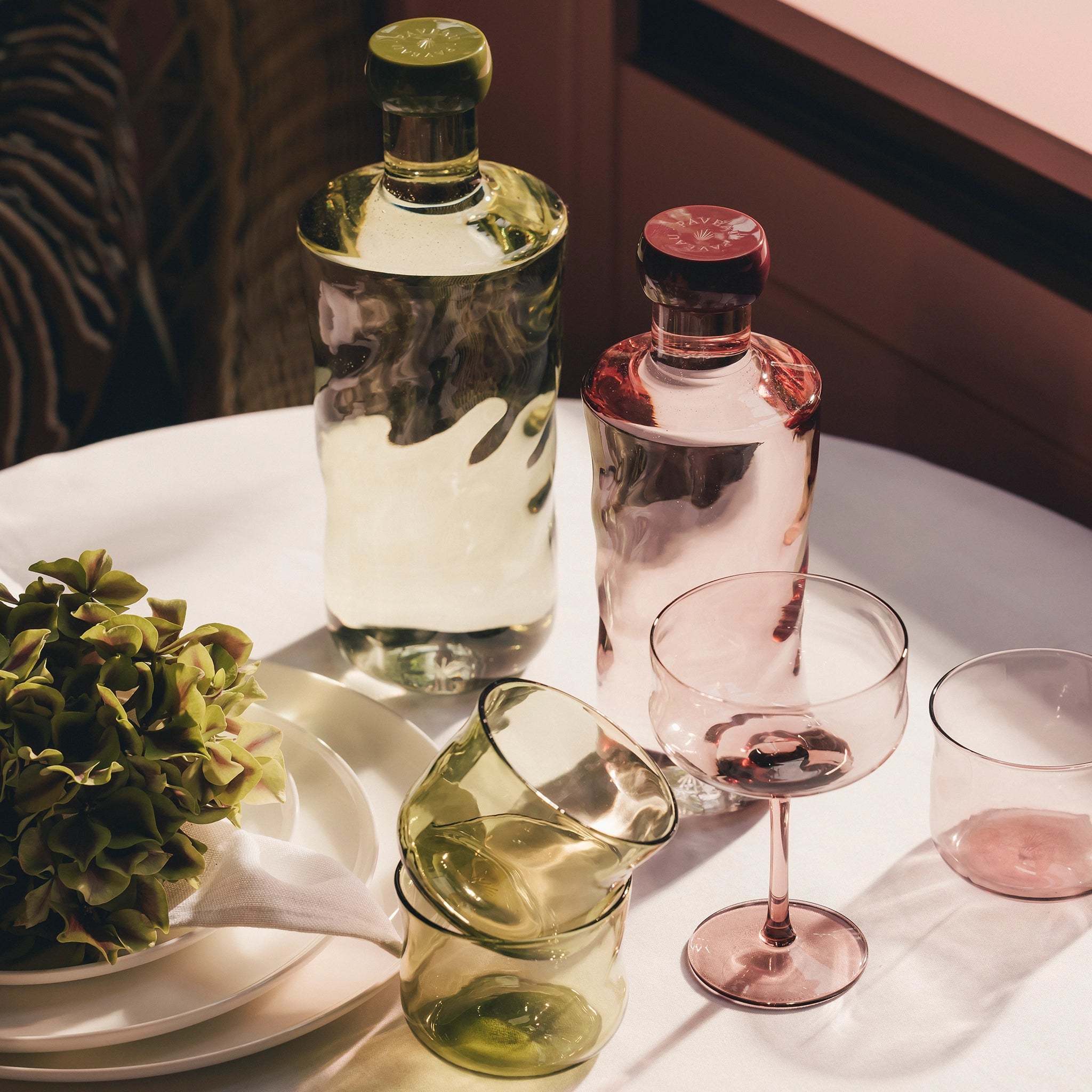 Glazen flessen, kopjes en een Paveau Swirl Waterglas Silt Bordeaux 24 cl van Paveau in groene en roze tinten staan op een wit tafelkleed naast borden met servetten en groene hortensia's, allemaal verlicht door zacht natuurlijk licht.