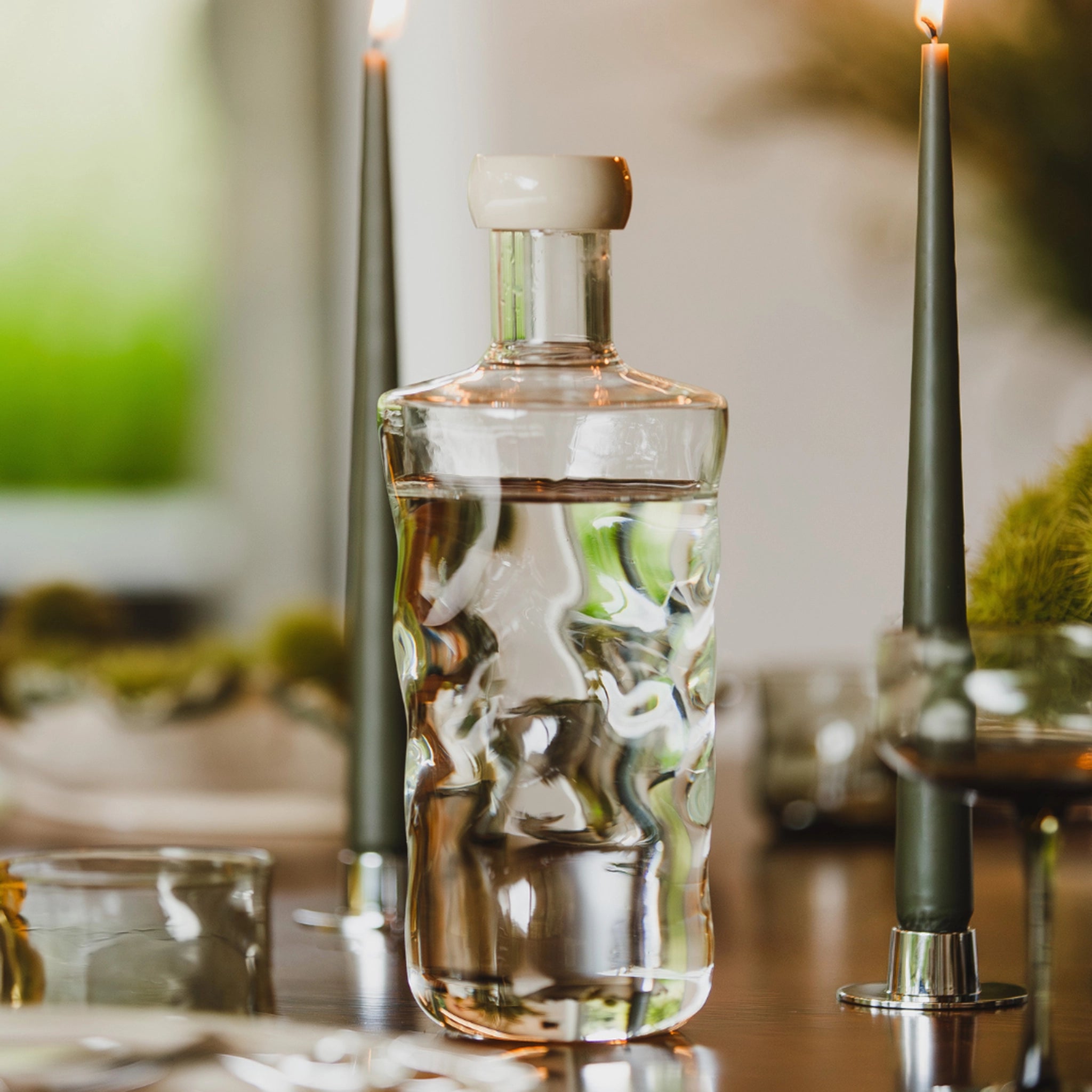 De Paveau Swirl Waterfles Wave Helder 0,75 l van Paveau met witte dop en textuur staat op een houten eettafel naast twee brandende kaarsen, met een vage groene achtergrond.