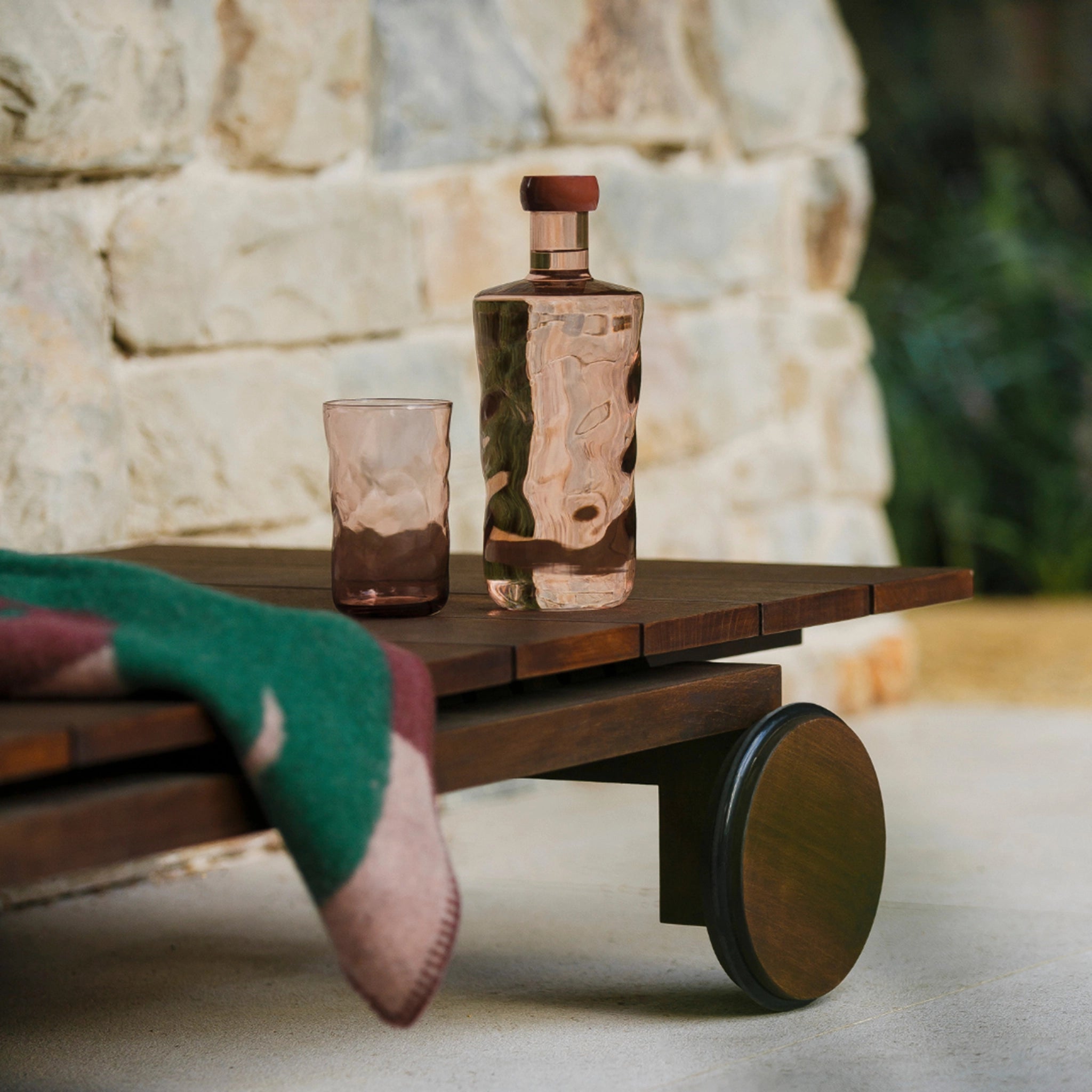 De Paveau Swirl Waterfles Silt Bordeaux 1,25 l van Paveau, gemaakt van elegant borosilicaatglas met een houten kap en bijpassend glas, staat op een houten bankje naast een deken en stenen muur - een stijlvol geschenk.