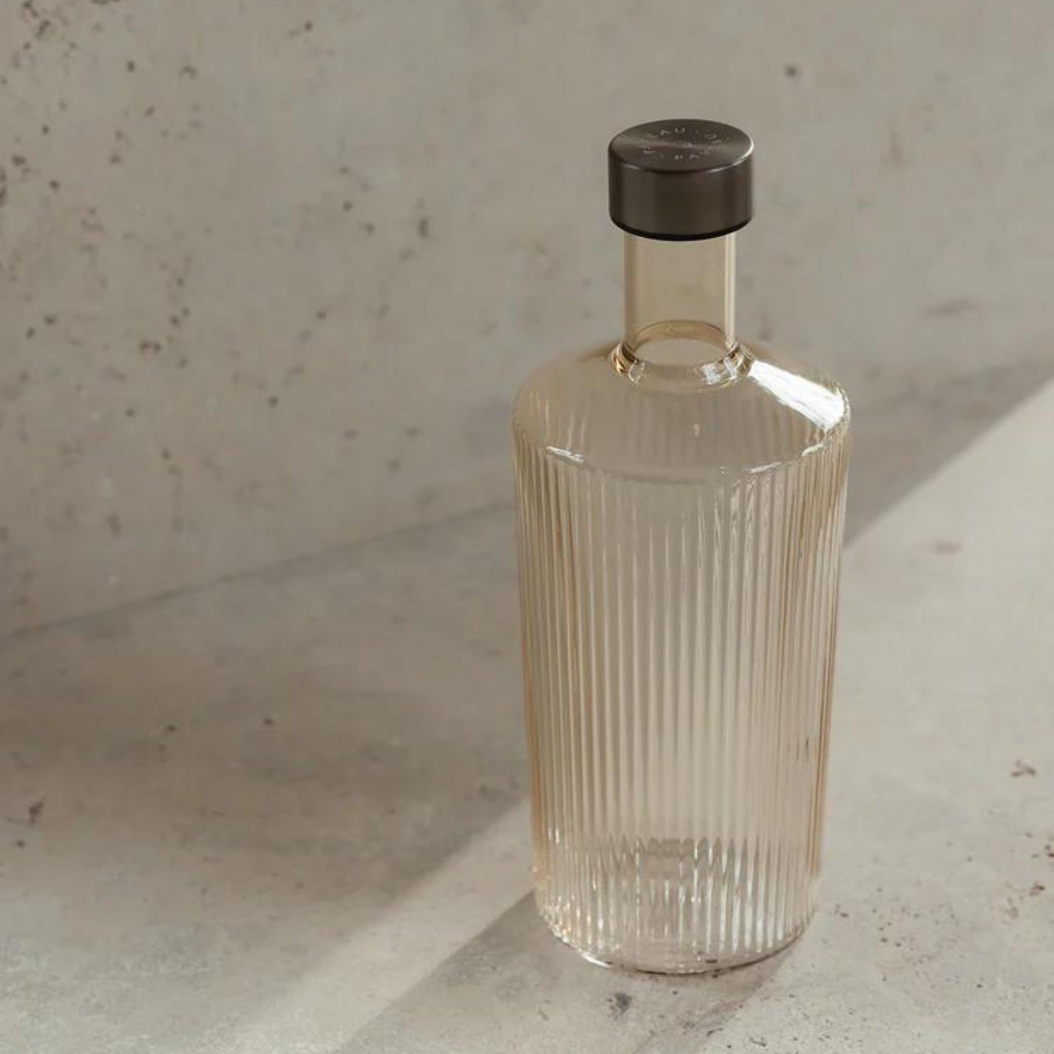 De Paveau Waterfles Bells Beige 1,25 l van Paveau heeft een helder, geribbeld ontwerp met een donkere kap, rustend op een licht stenen oppervlak. Zonlicht versterkt de minimalistische en elegante stijl.