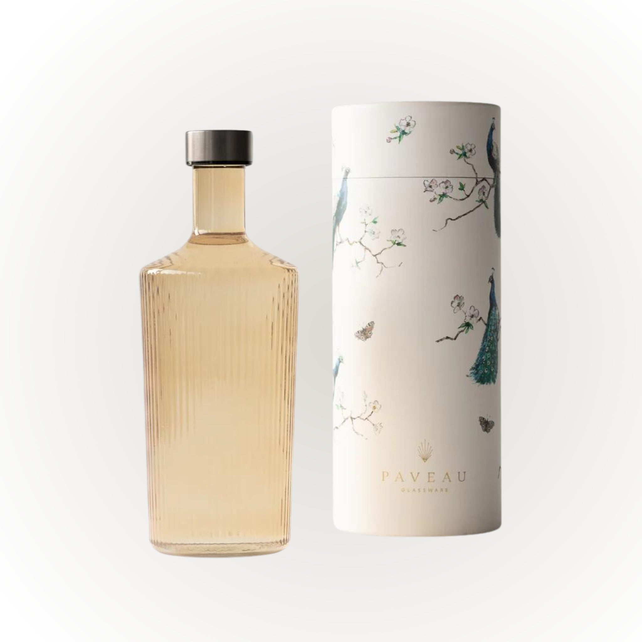 Een hoge, geribbelde Paveau Waterfles Bells Beige 1.25L met een donkere kap staat naast een cilindrische witte doos versierd met pauwen, takken en vlinders, met het label "PAVEAU GASTRONOME" in goud.