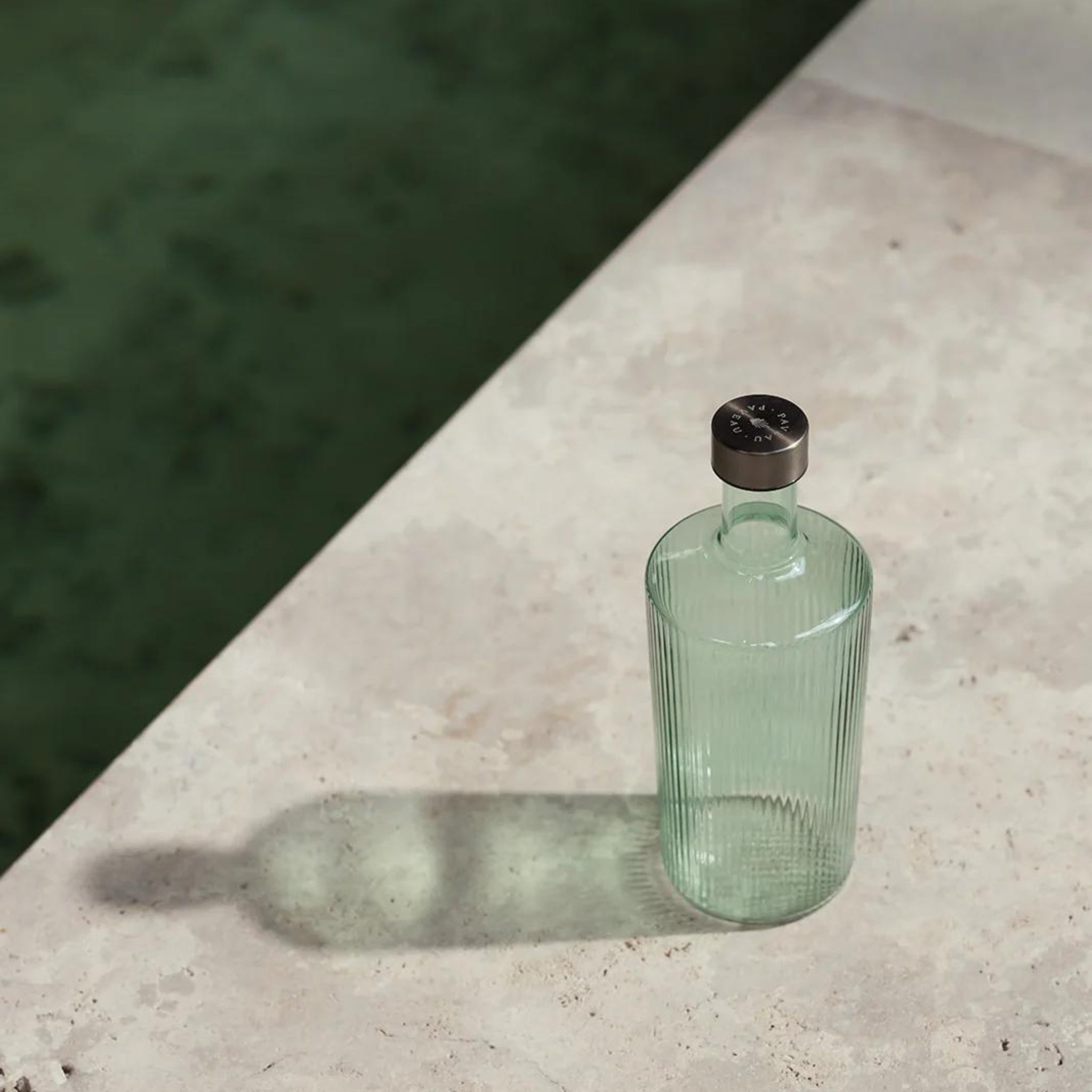 De Paveau Waterfles Bondi Green 1,25 l van Paveau, een heldere geribbelde fles met een donkere dop, werpt zijn schaduw op een licht stenen oppervlak bij groen water.