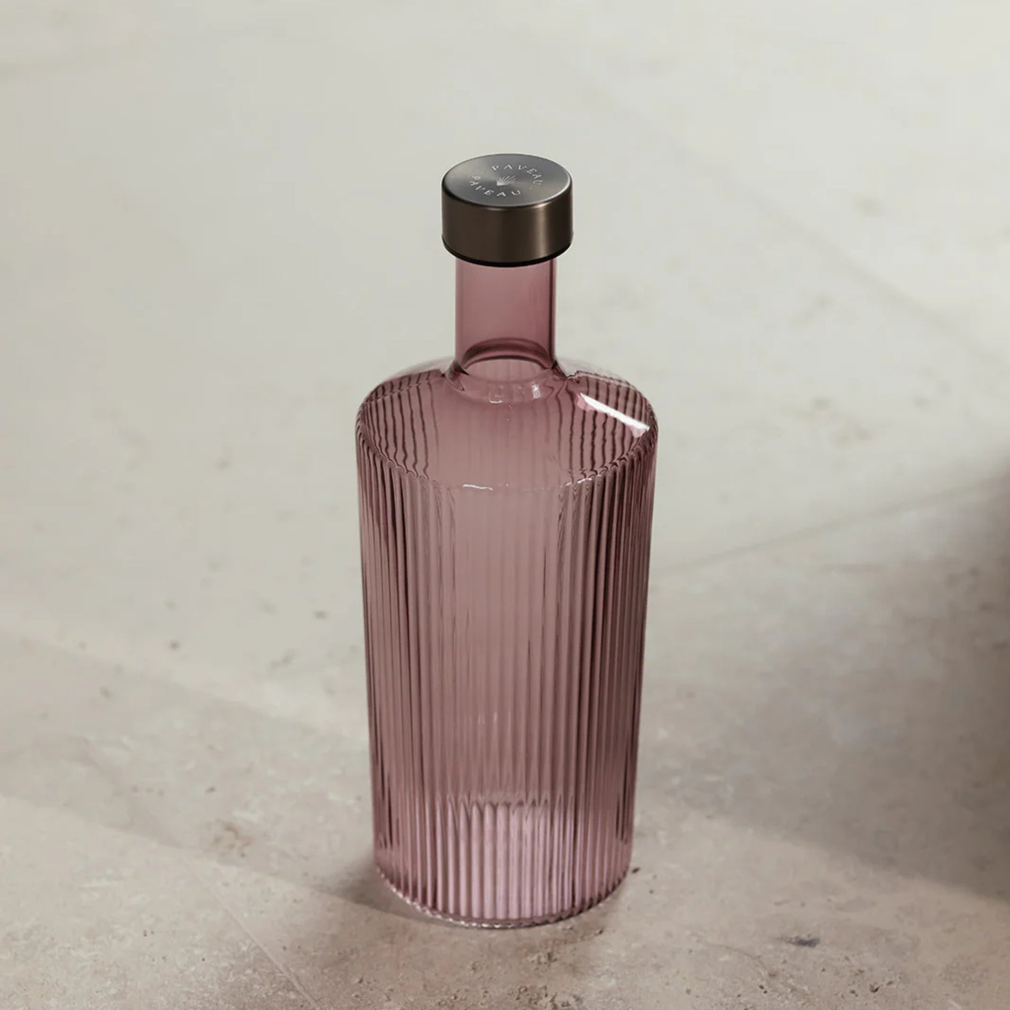 De Paveau Waterfles Broome 1,25 l van Paveau heeft een hoog, geribbeld geglazuurd ontwerp in stoffig roze met een metalen kap, weergegeven op een licht stenen oppervlak.