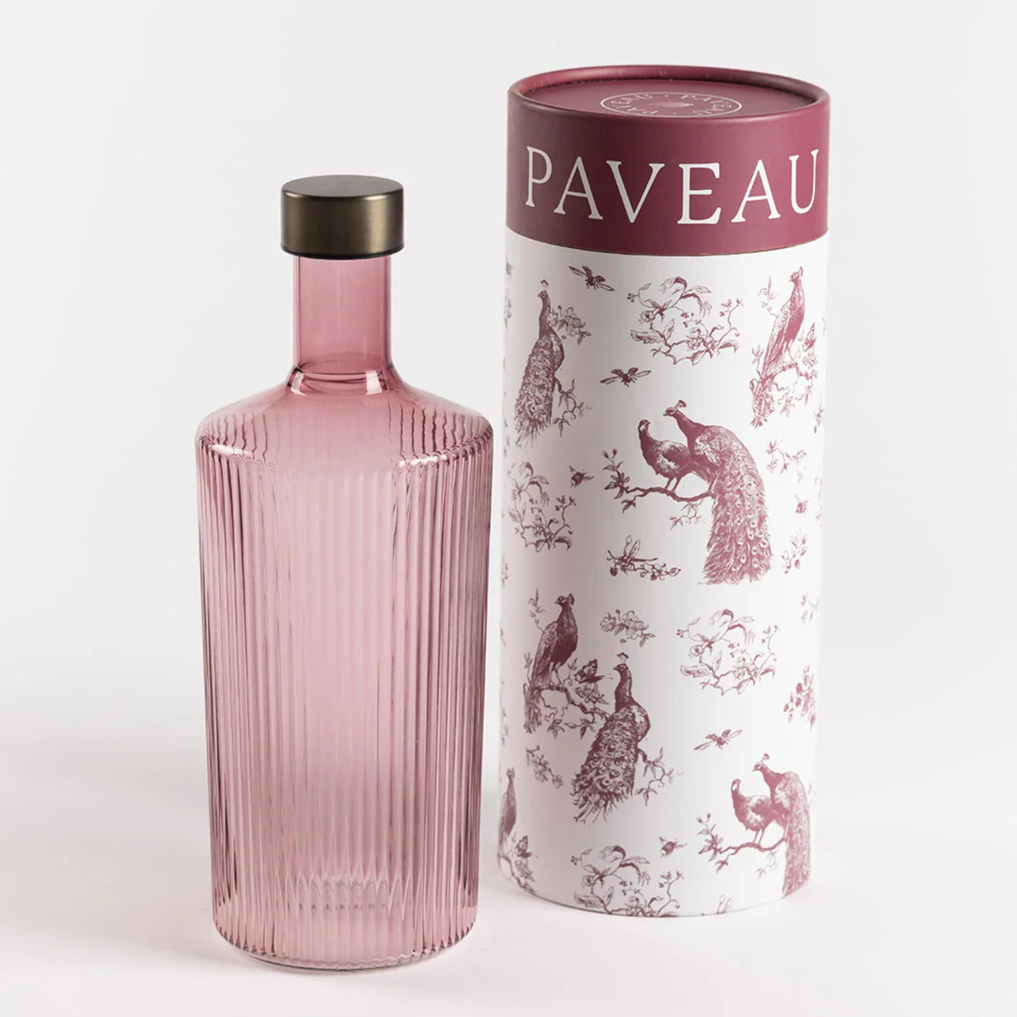 Een geribbelde, roze glazen Paveau Waterfles Broome 1,25 l met een donkere dop staat naast een witte cilindrische doos versierd met bordeauxrode pauwen en bomen, met "PAVEAU" bovenaan.