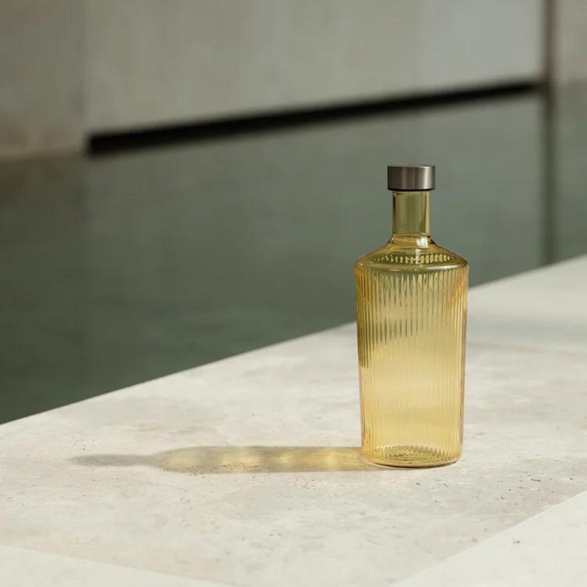 De Paveau Waterfles Cable Yellow 1,25 l van Paveau, met een geribbeld amberkleurig design en metalen kap, rust op een licht stenen oppervlak met een lange schaduw; water en neutrale muren vervagen op de achtergrond.