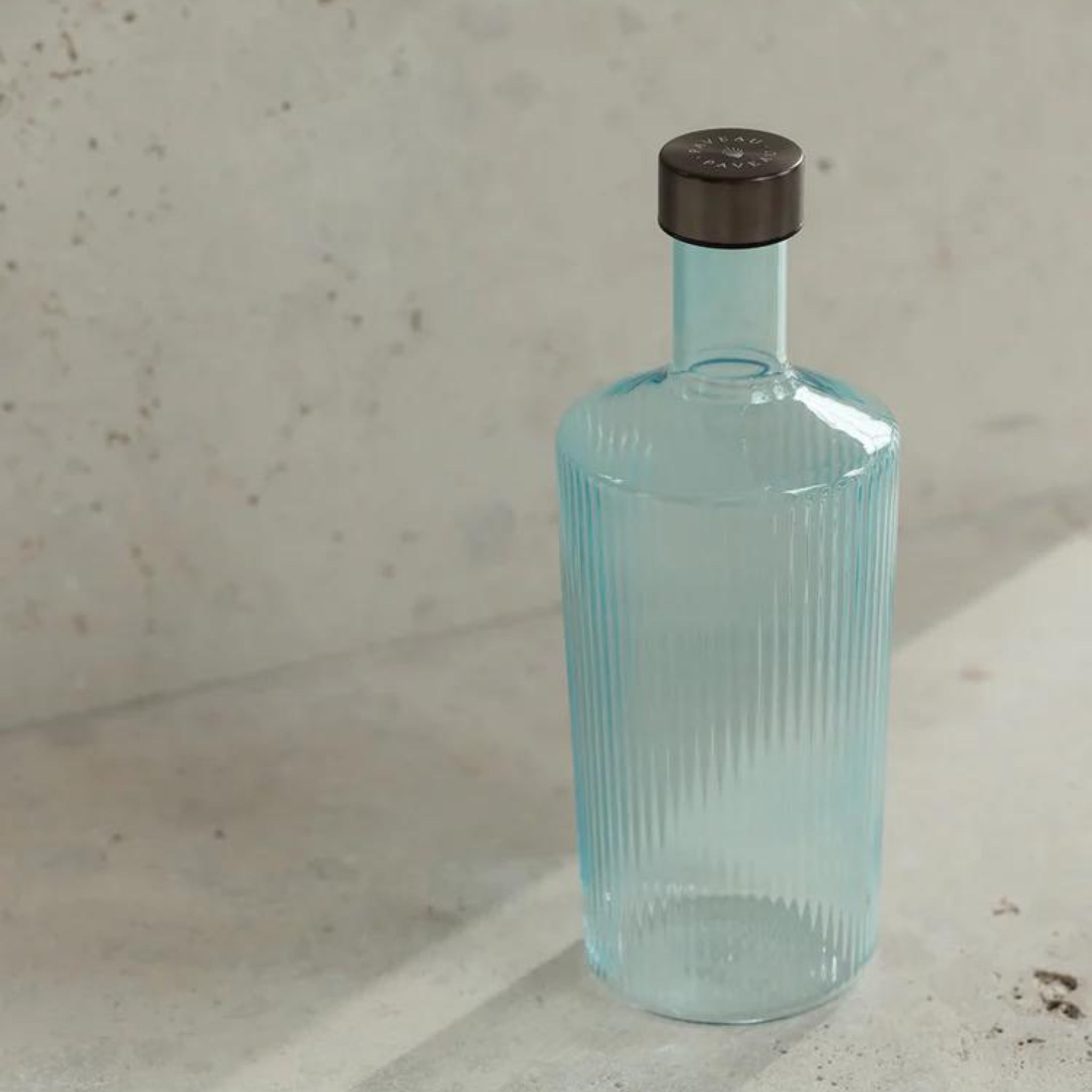 De Paveau Waterfles Lucky Turquoise 1,25 l van Paveau is een hoge, geribbelde, lichtblauwe glazen fles met een donkere dop op een licht, gestructureerd oppervlak tegen een neutrale muur.