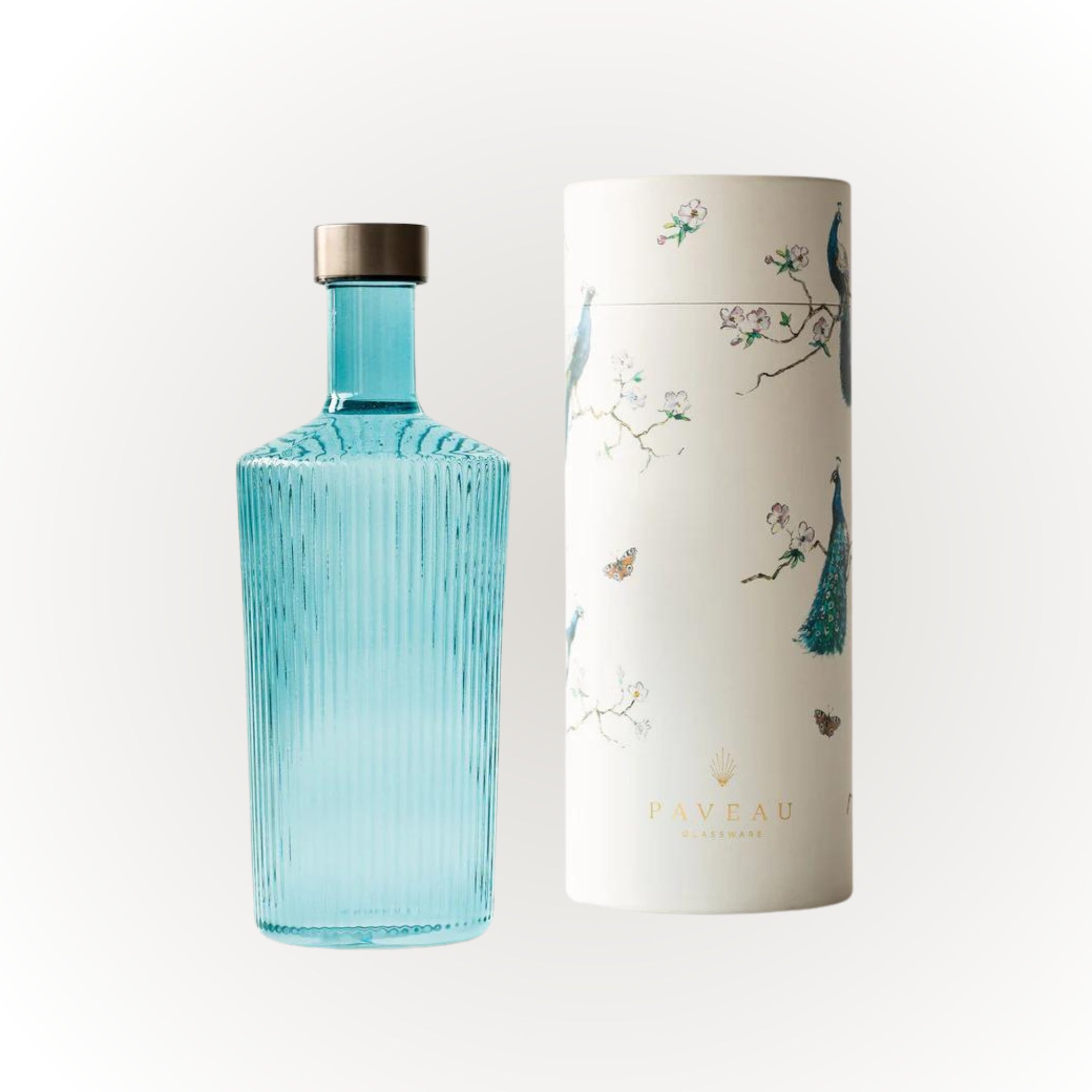 Een Paveau Waterfles Lucky Turquoise 1,25 l met een metalen dop staat naast een hoge witte cilindrische doos versierd met bloemmotieven, vlinders en pauwen. Op de bodem van de doos staat het merk Paveau.