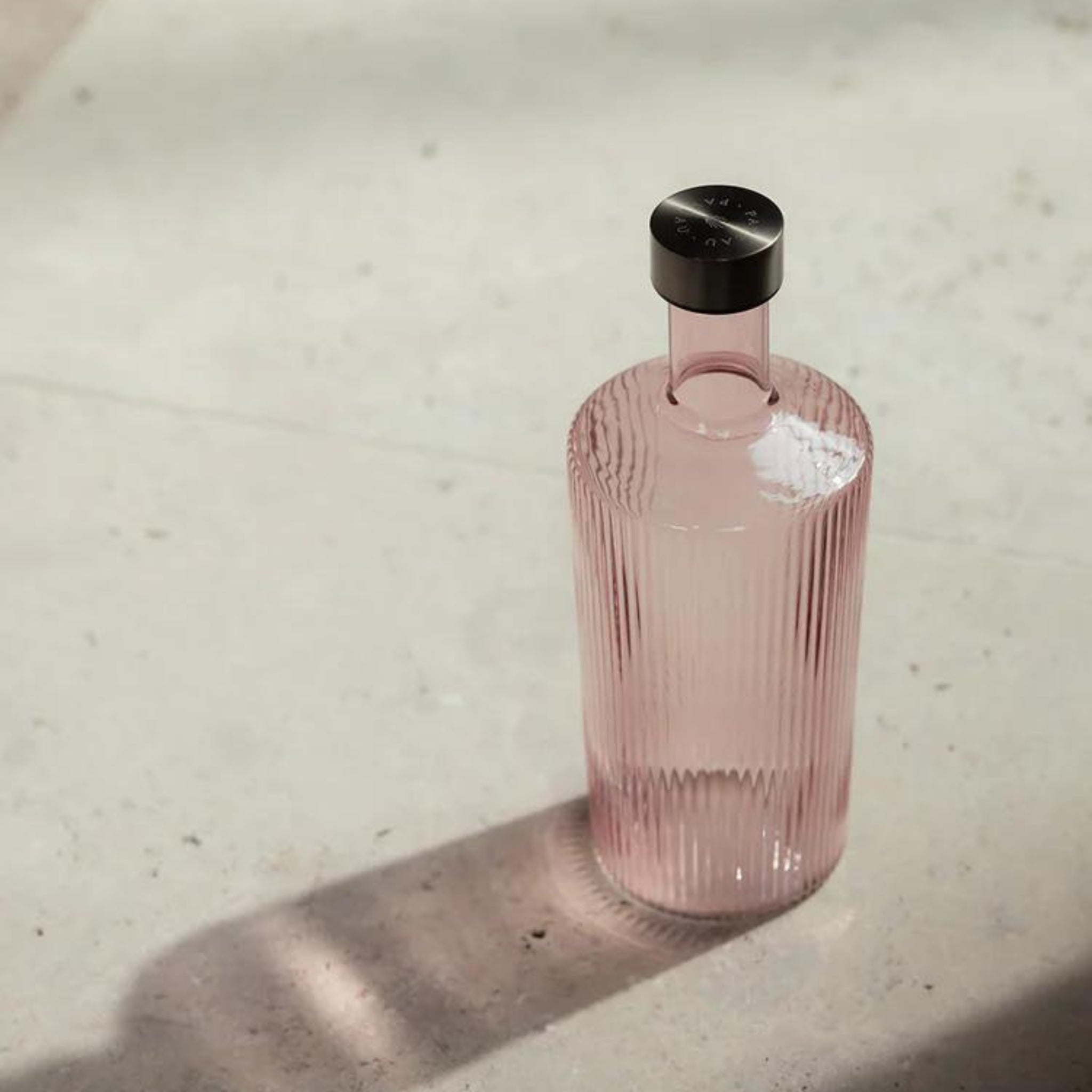 De Paveau Waterfles Pink 1,25 l van Paveau, met een geribbeld doorschijnend roze ontwerp en metalen dop, staat op een licht oppervlak met een zachte schaduw in natuurlijk zonlicht.