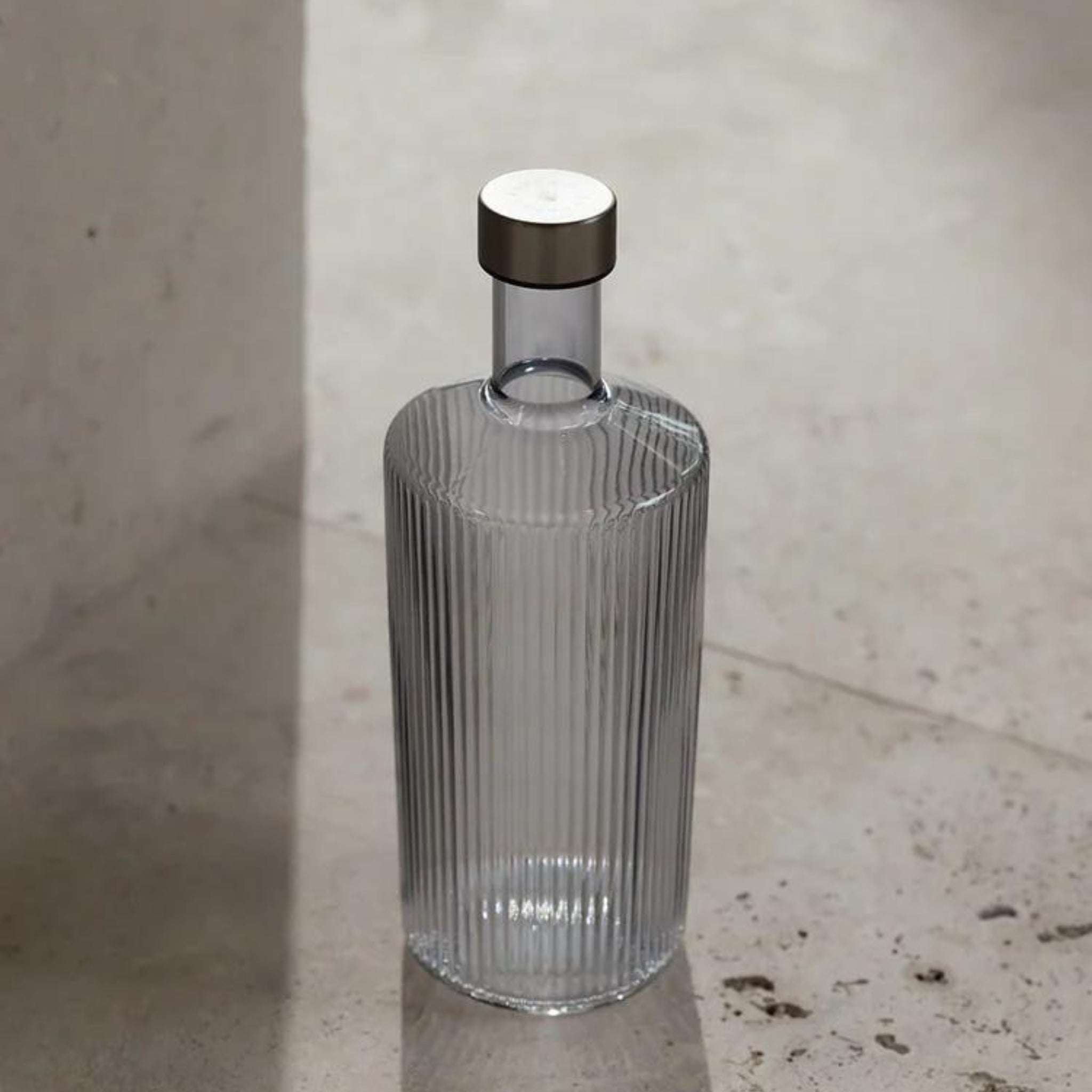 De Paveau Waterfles Twilight Blue 1,25 l van Paveau, met een helder geribbeld ontwerp en metalen kap, staat rechtop op een beige stenen oppervlak dat baadt in zacht natuurlijk licht.