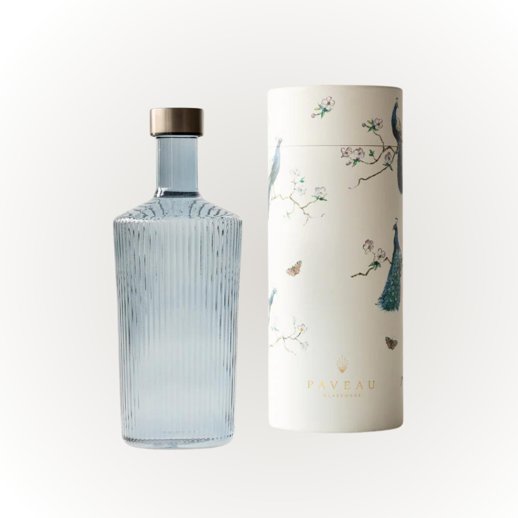 Een heldere, geribbelde Paveau Waterfles Twilight Blue 1,25 l met een metalen dop staat naast een hoge witte cilinderkoffer van Paveau, versierd met pauwen, bloemen en vlinders.